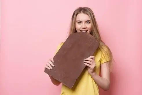 A woman taking a bite out of an oversized chocolate bar, against a pink backdrop.