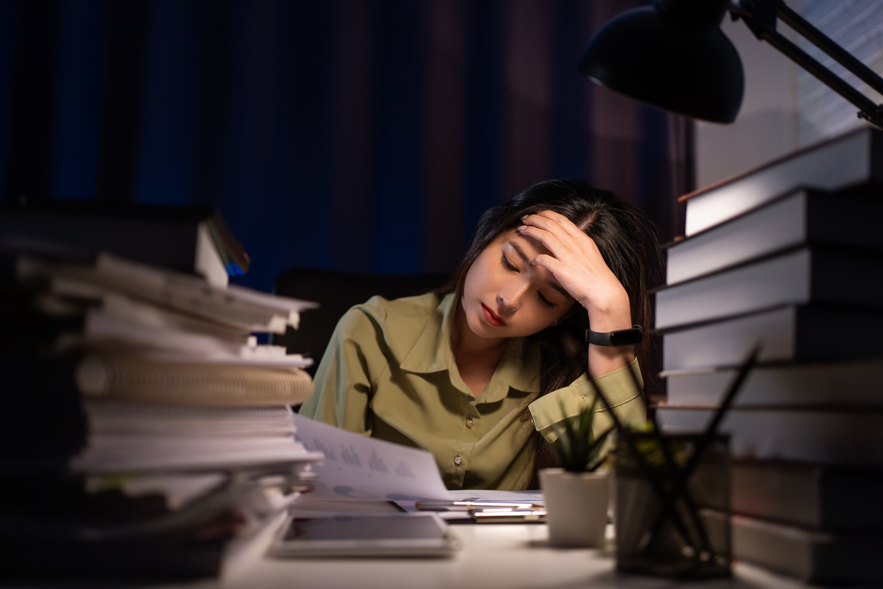 A woman looks at a sheet of paper with charts on it, under a table light in a darkened room, with stacks of folders and books on the table in the foreground.
