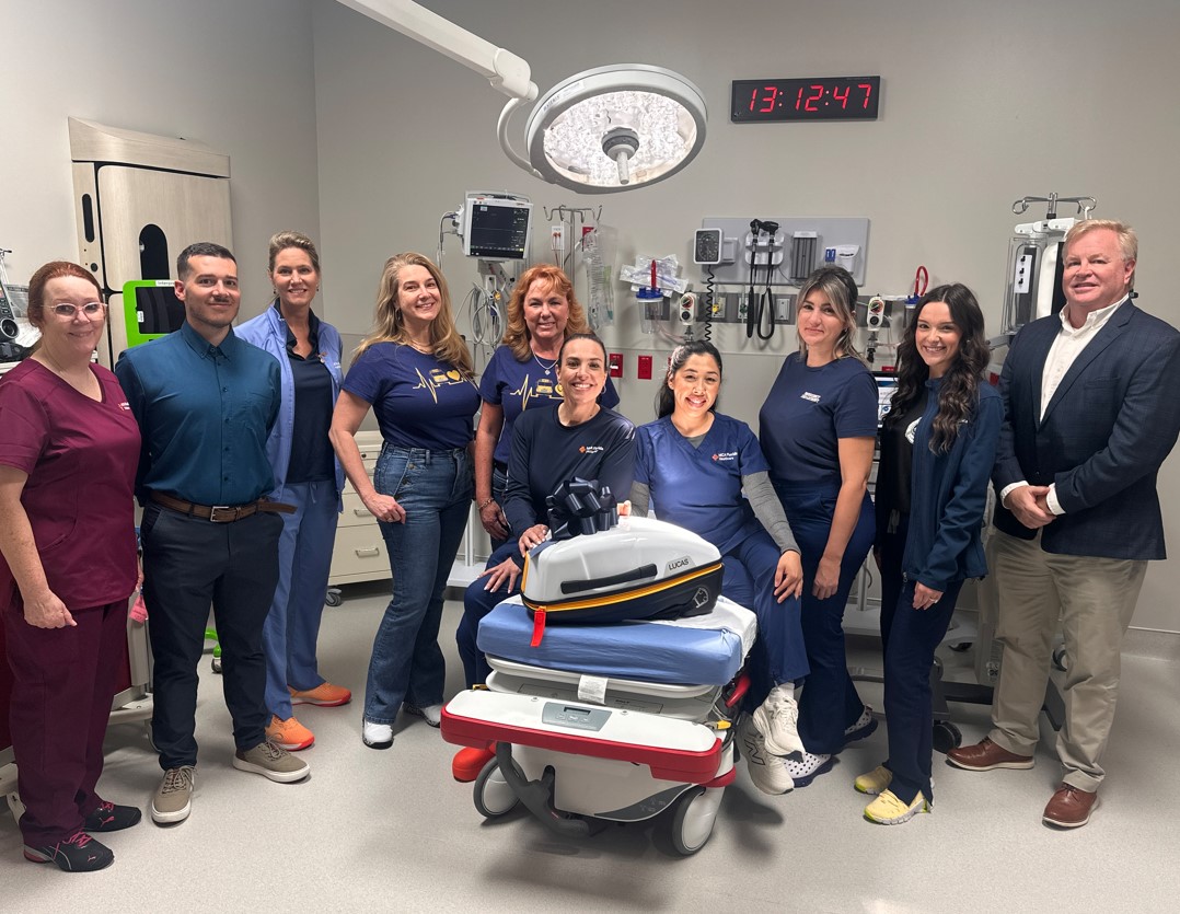 A group of people stands in an emergency room surrounded by medical equipment, including monitors, IV stands and a mobile multi-compartment device. Some individuals wear scrubs, while others are dressed in casual or business attire. A digital wall clock reads 13:12:47, indicating the time of the gathering. The setting is an emergency triage room.