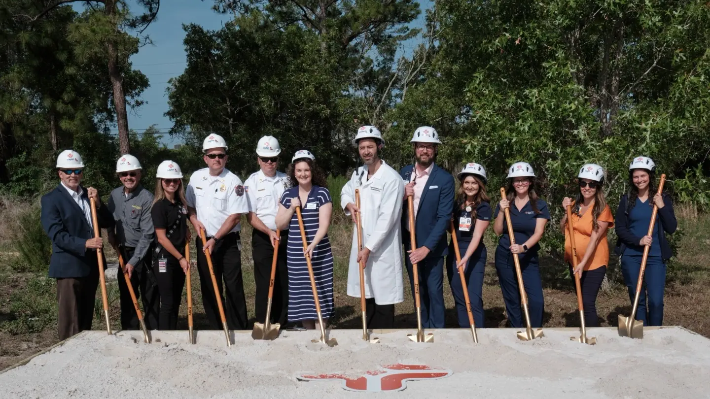 Group of individuals with construction hats holding shovels in the sand surrounding the HCA logo.