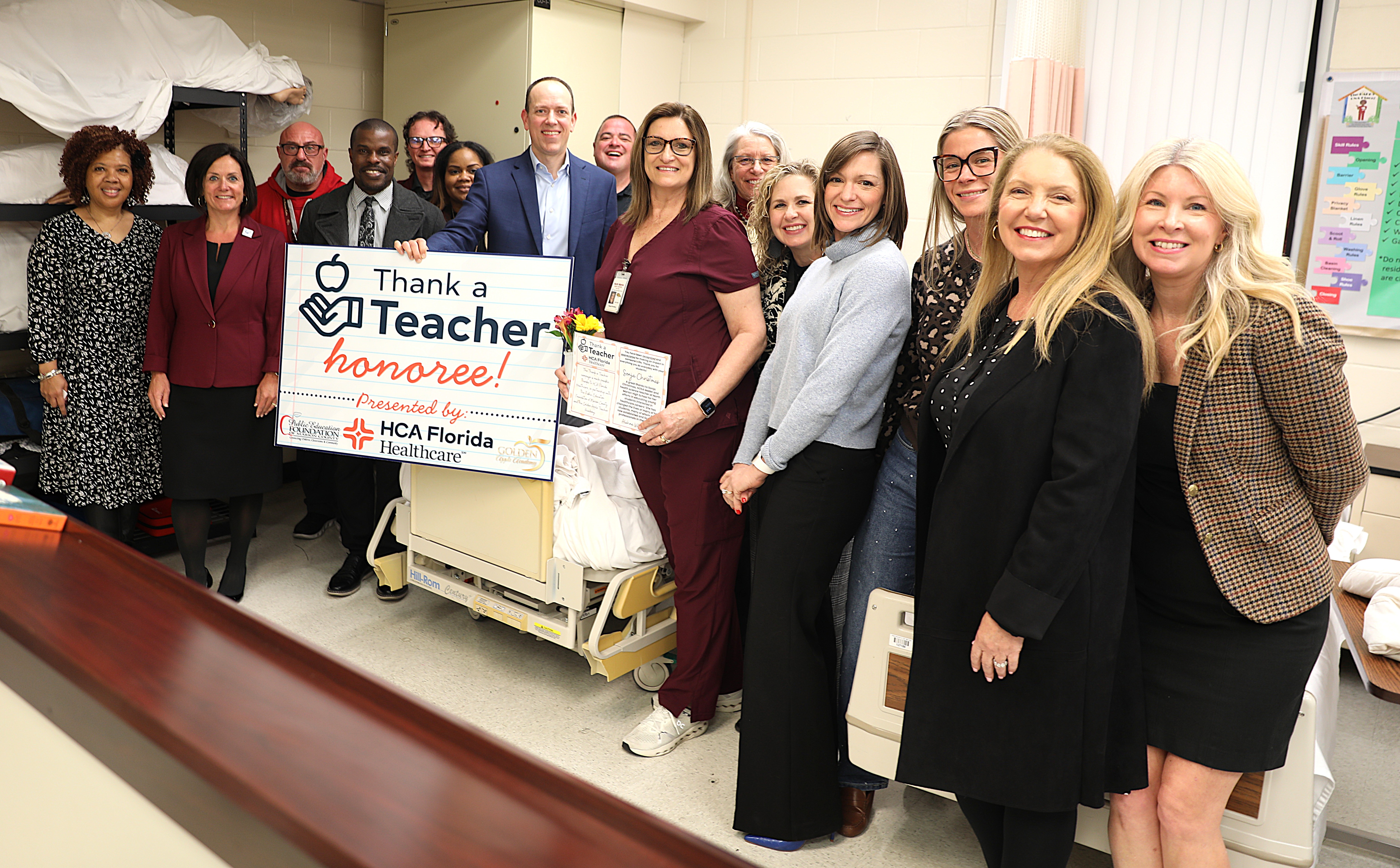 A group of educators and healthcare professionals gather in a clinical training classroom around a hospital bed, smiling for a photo. Several people hold a large "Thank a Teacher Honoree – Presented by HCA Florida Healthcare" sign while the honoree holds flowers and a certificate.