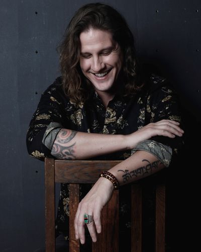 Eric Breish sits backwards on a wooden chair, smiling and gazing downward.