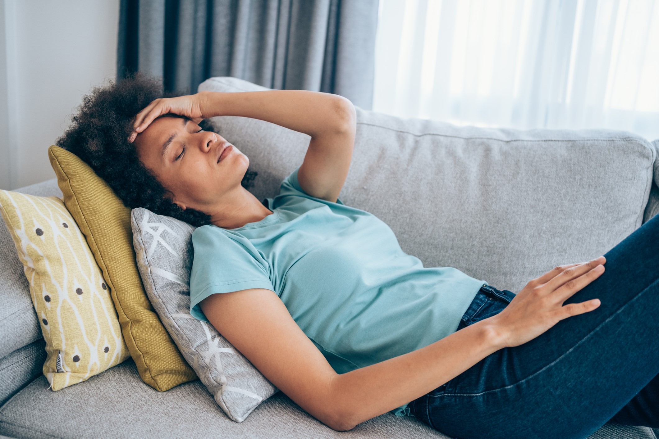 A woman reclining on a couch, with her eyes closed and her hand on her head, in apparent discomfort.