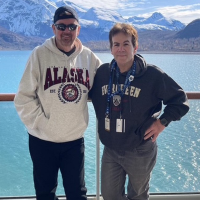 Two men in hoodies stand side by side on a boat deck, one wearing an "Alaska" sweatshirt and the other an "Evergreen" sweatshirt, with bright blue water and snow‑capped mountains in the background.