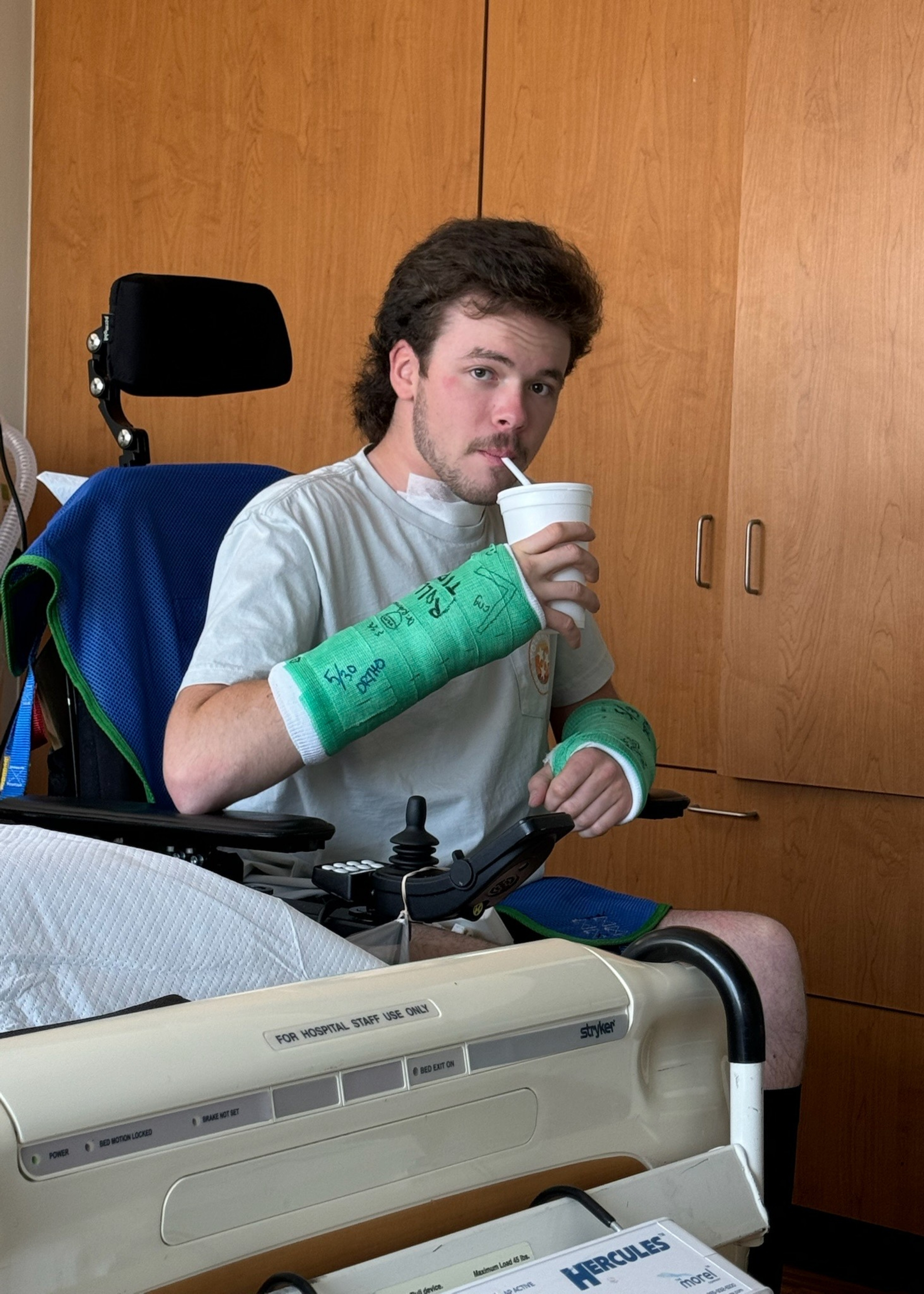 Bo Bryant in wheelchair with wrist casts in hospital room drinking from cup