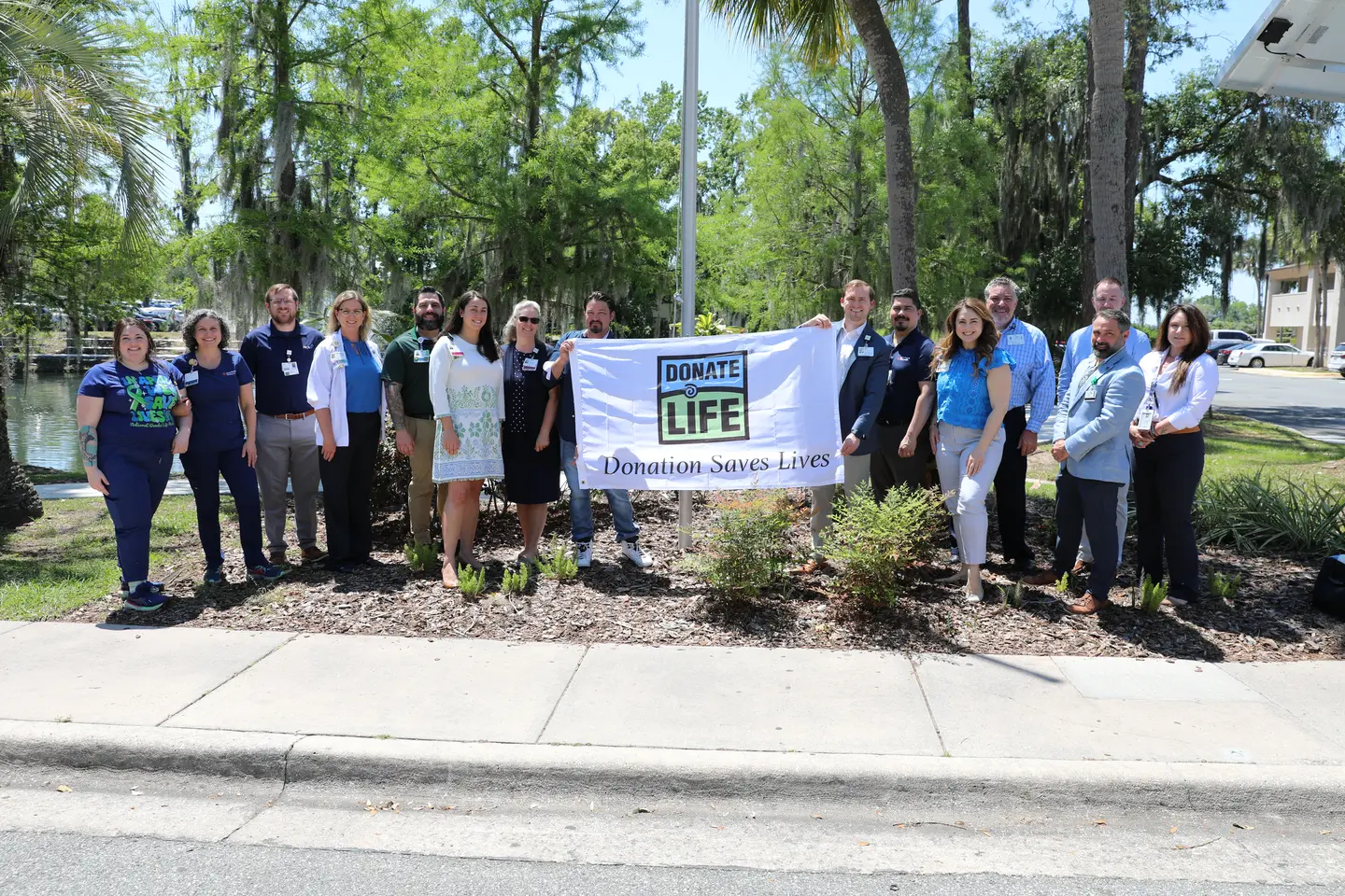 Photo shows a group of people outside under a flagpole, smiling and holding a white flag that says "Donate Life, Donation Saves Lives".