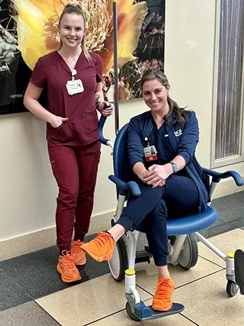 A female nurse standing next to a female nurse sitting in a wheelchair smiling.