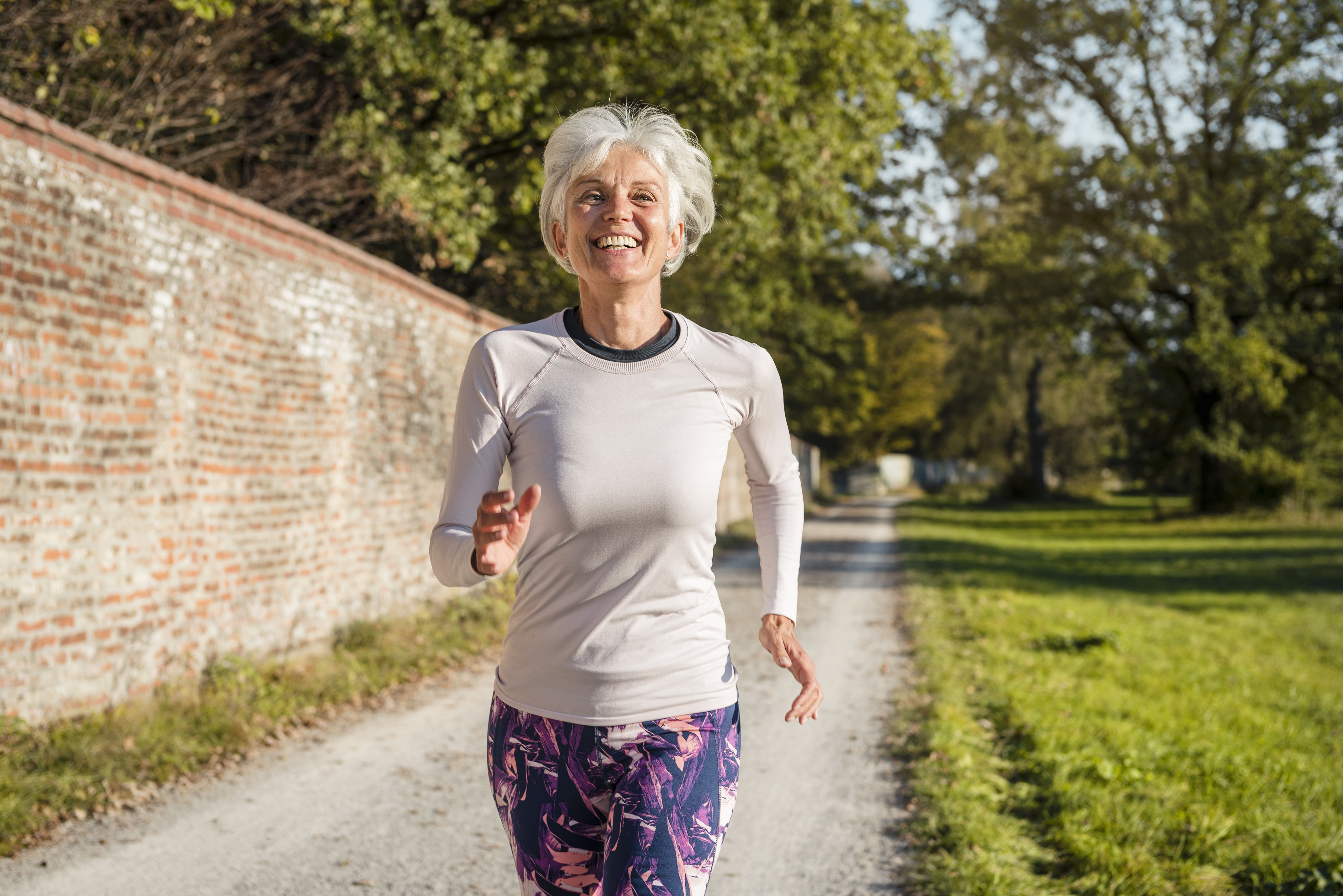 A smiling senior woman jogging outdoors on a dirt path, alongside a brick wall in a park.