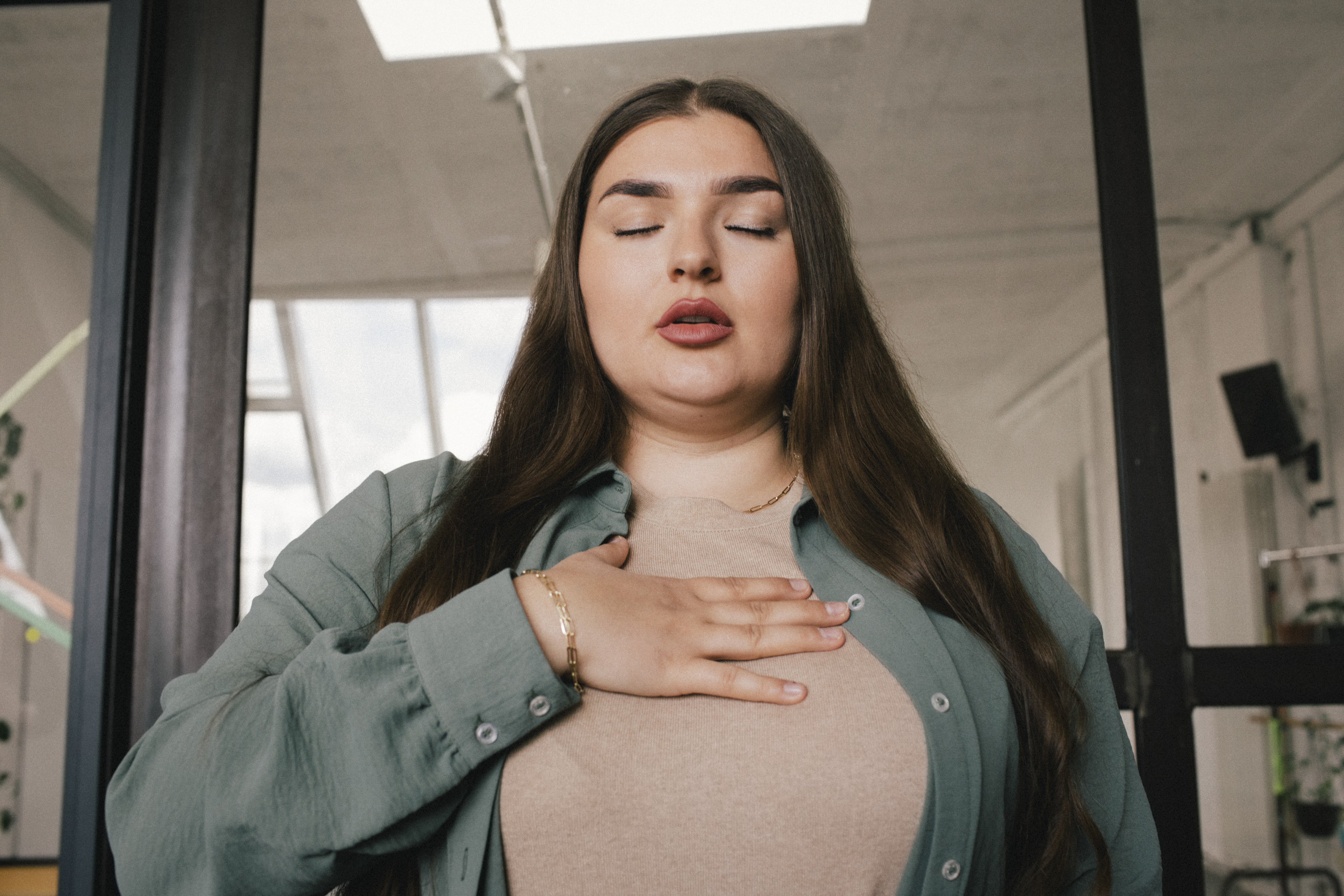 A young woman with her eyes closed and her hand on her chest, while standing in the office.