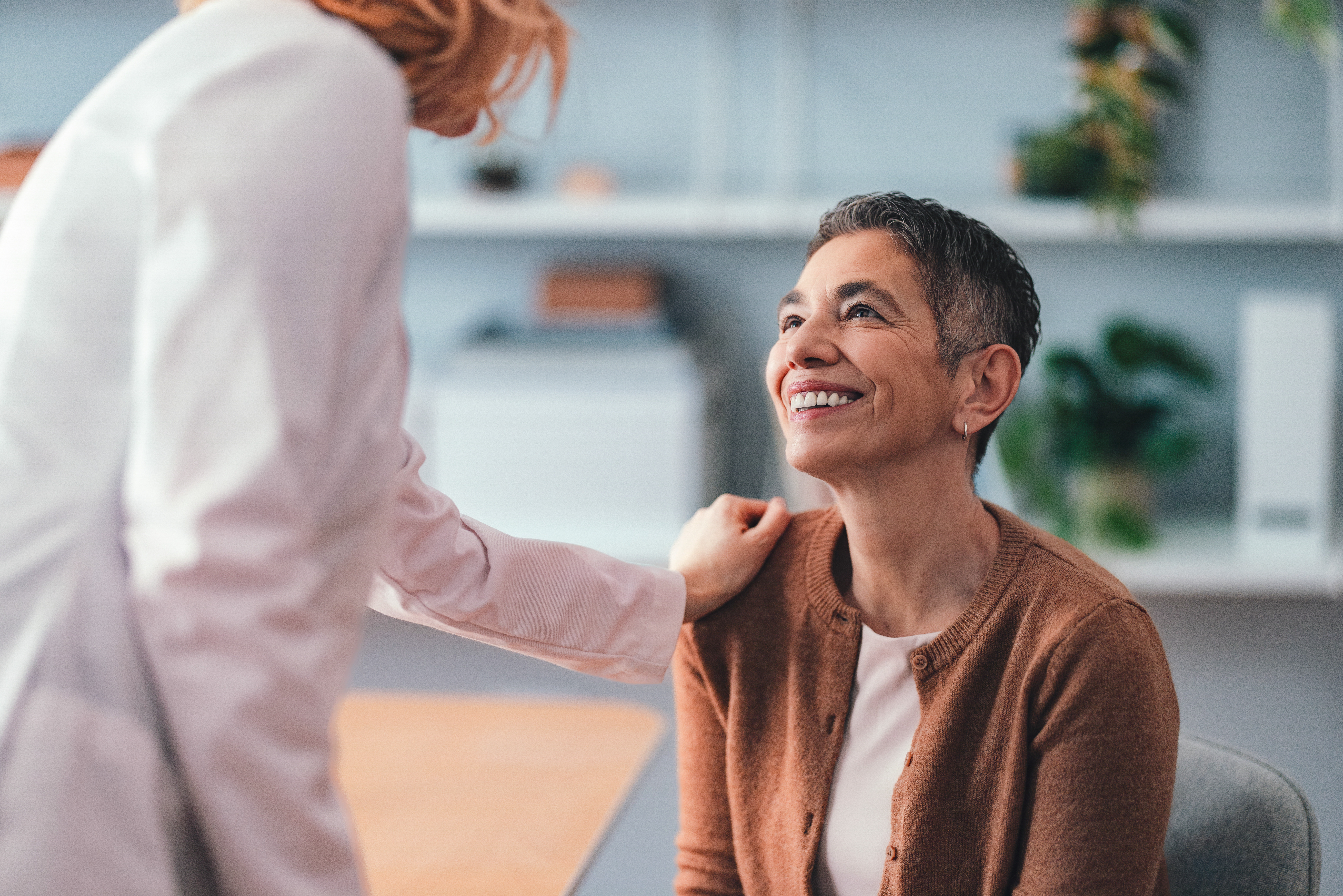 A doctor places her hand on a smiling patient's shoulder, in an office.