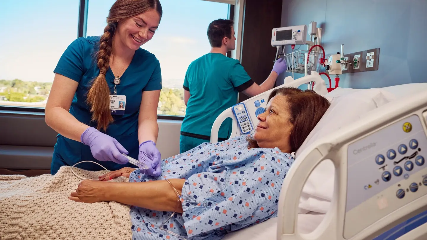 Nurse administering medicine through a patient's IV while they lay in a hospital bed.