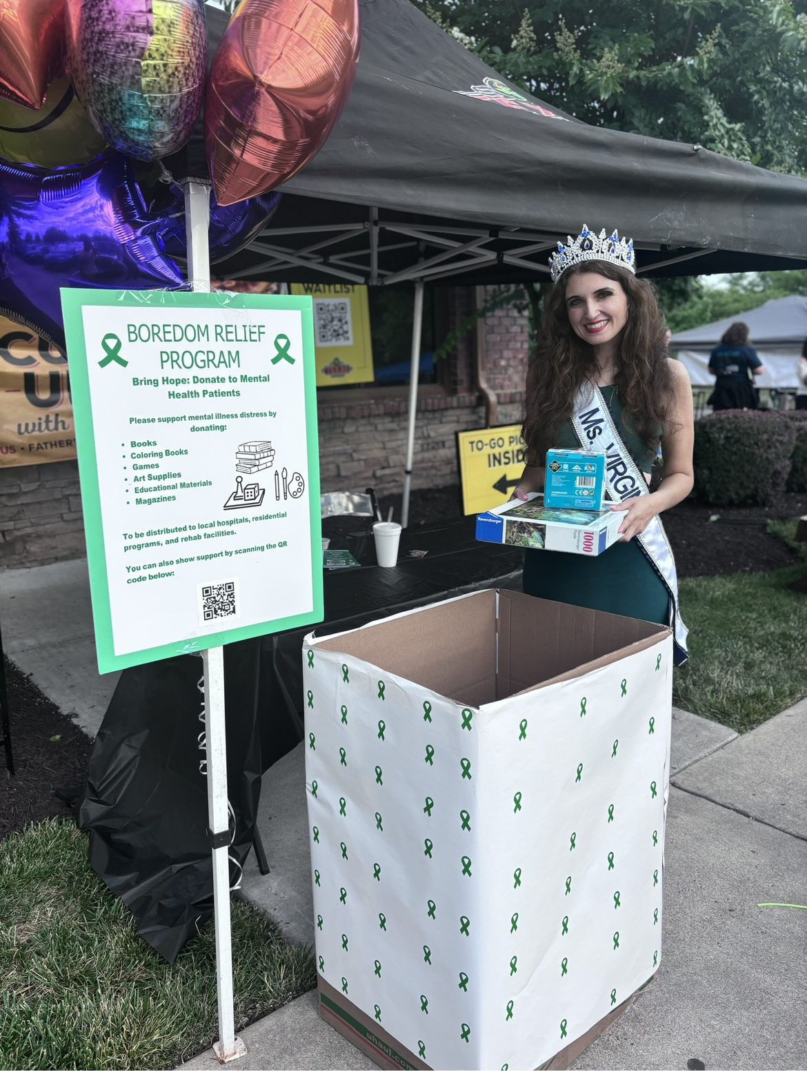 Taylor Ragano, in Miss Virginia sash and crown, placing puzzles and books into Boredom Relief Program donation box.