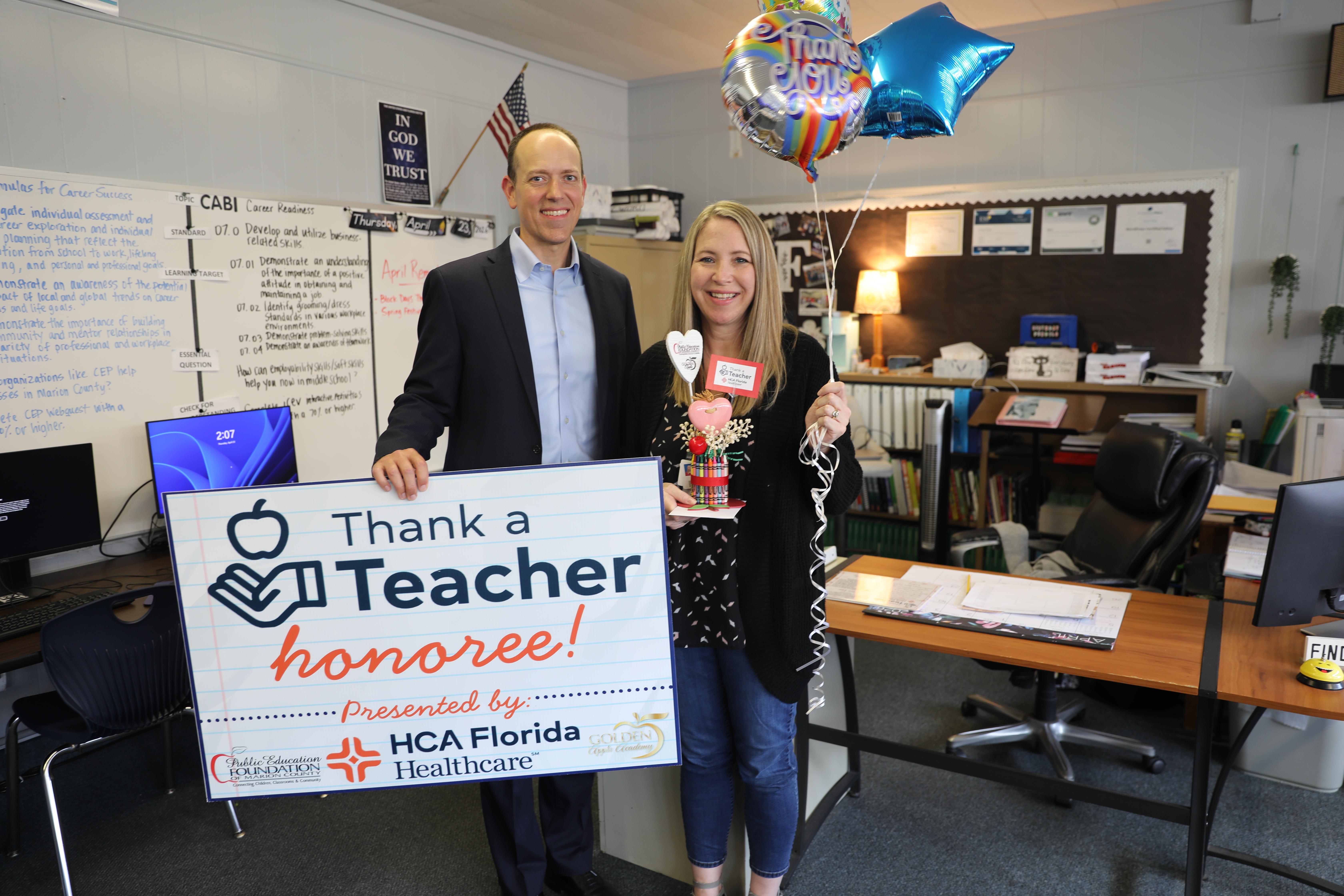 A man and a female teacher stand together in a classroom holding a large sign that reads "Thank a Teacher honoree!" The teacher holds balloons and a small gift, and both are smiling. A desk, computer, and classroom posters are visible behind them.