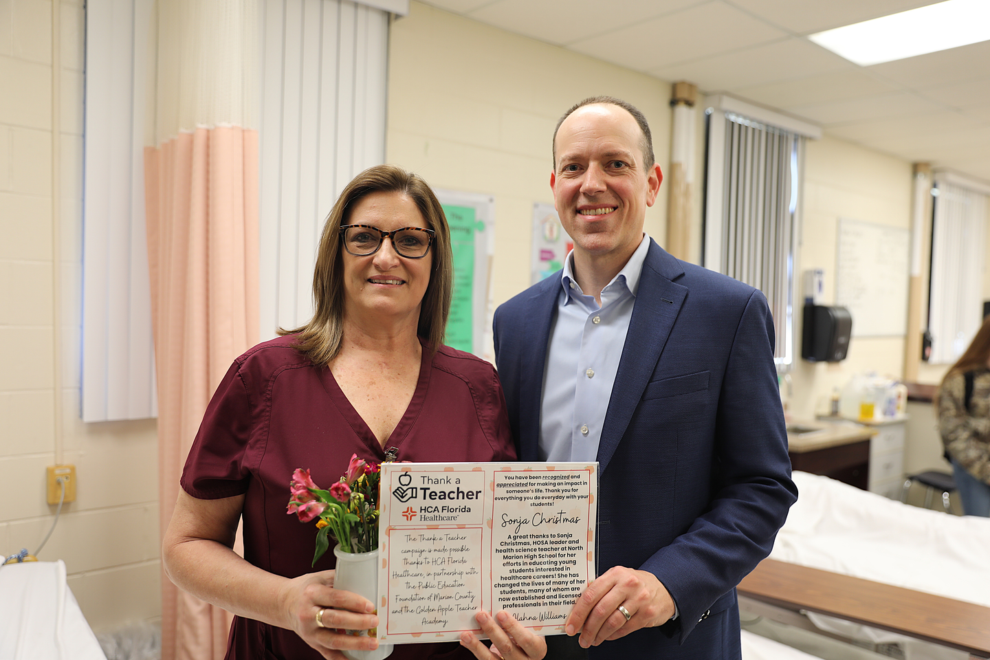 A woman in maroon scrubs and a man in a blue suit stand side by side in a healthcare classroom, smiling at the camera while holding a "Thank a Teacher – HCA Florida Healthcare" certificate and flowers. A hospital bed and clinical training supplies are visible behind them.