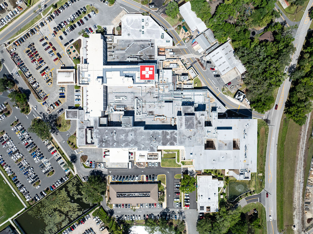 Aerial view of HCA Florida Ocala Hospital showing the full hospital complex, surrounding parking lots filled with cars, nearby roads, and a rooftop helipad marked with a red "H."