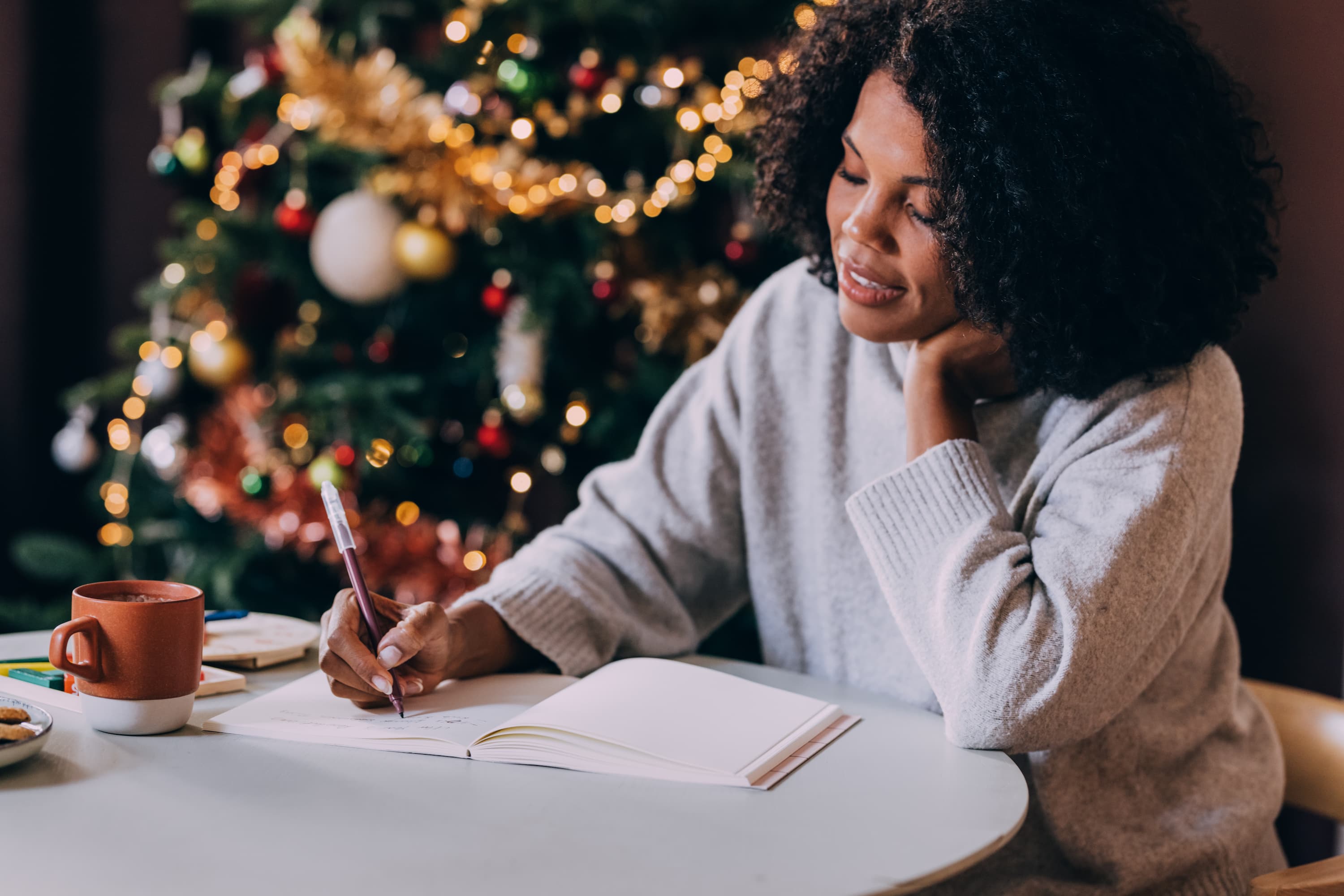 A woman writes in a journal while seated at a table next to a Christmas tree.
