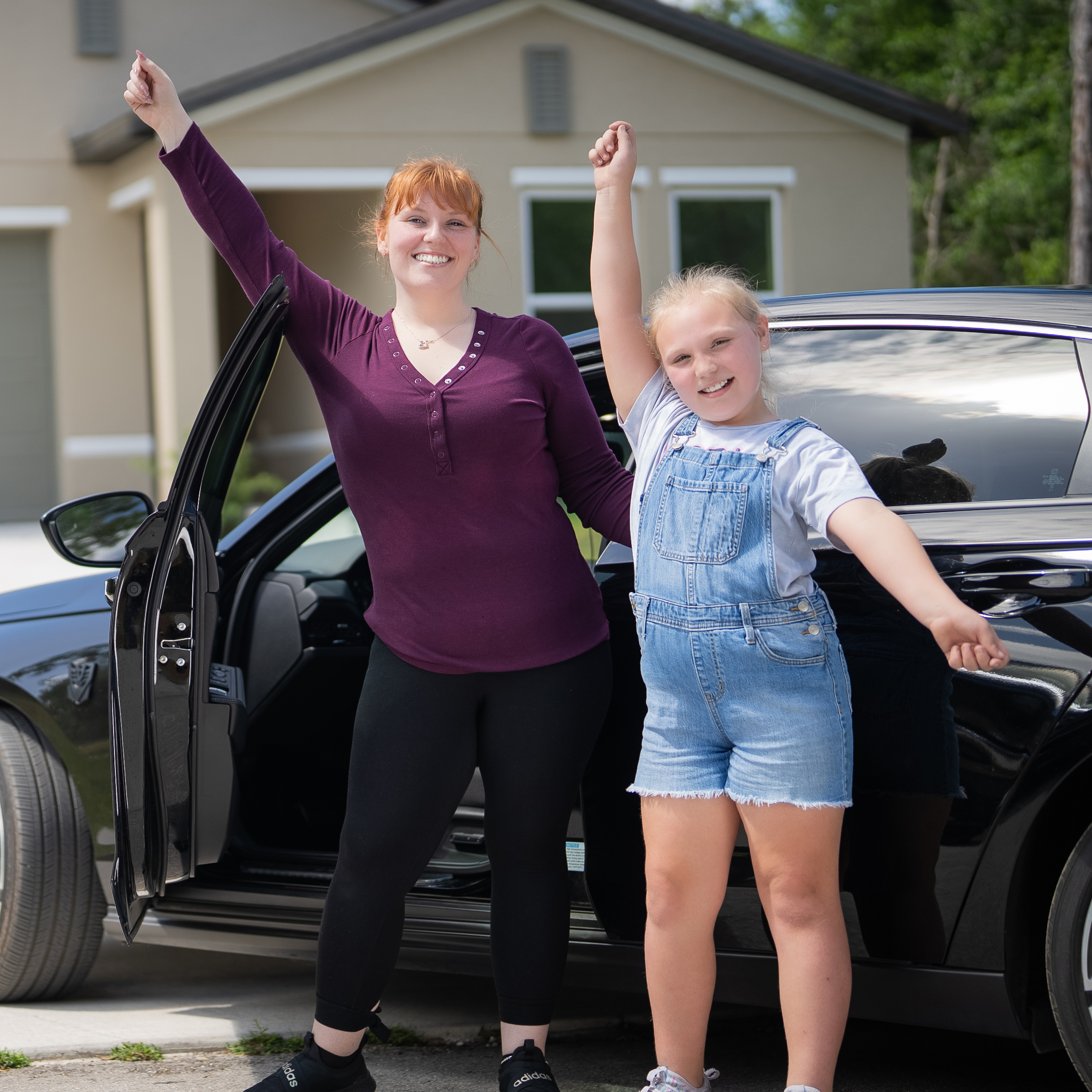 A woman and her young daughter are smiling and each holding one hand in the air to celebrate as they stand in front of a black car.