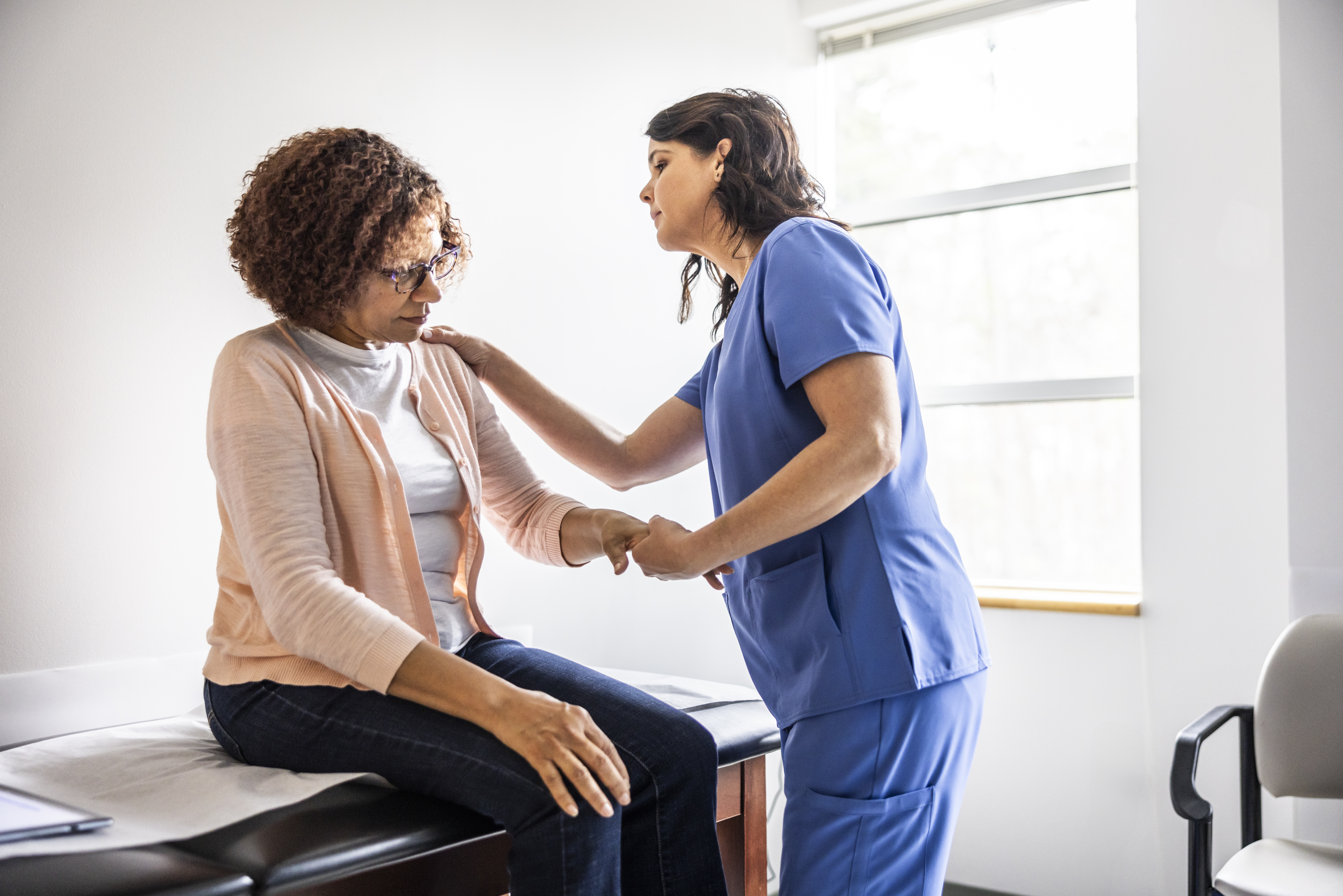 A healthcare professional in blue scrubs steadies a seated woman, during an exam in a bright exam room.