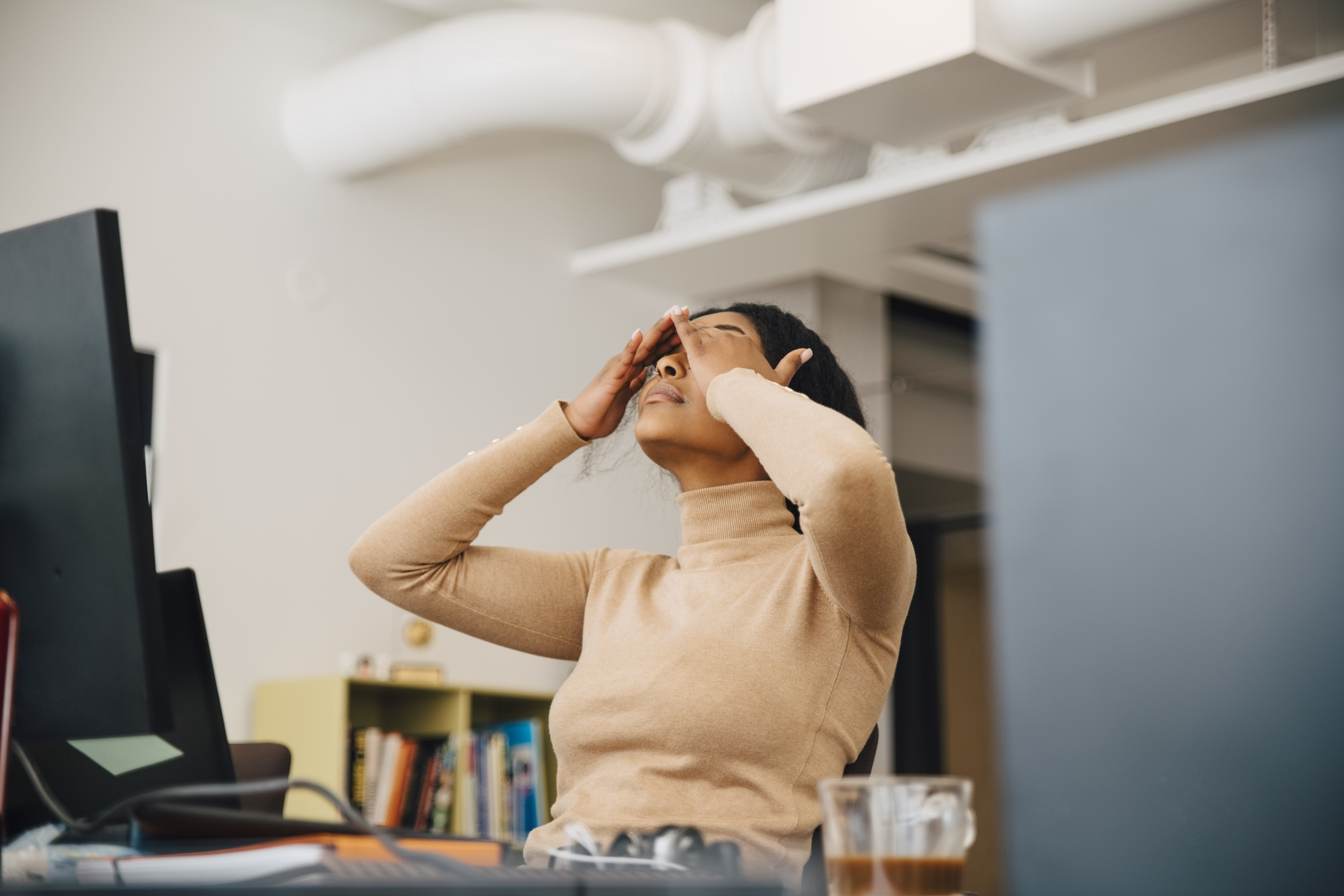 A woman holds her head in her hands while leaning back in her desk chair at work.