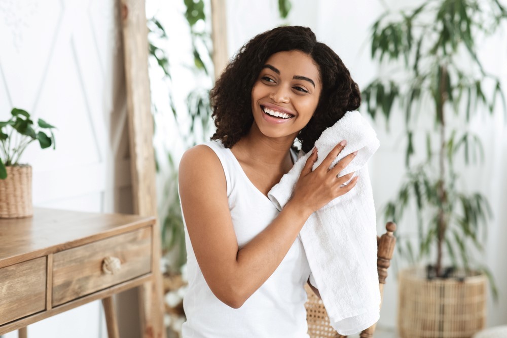 A woman drying her hair with a towel, in a bedroom with a dresser, mirror, and plants.