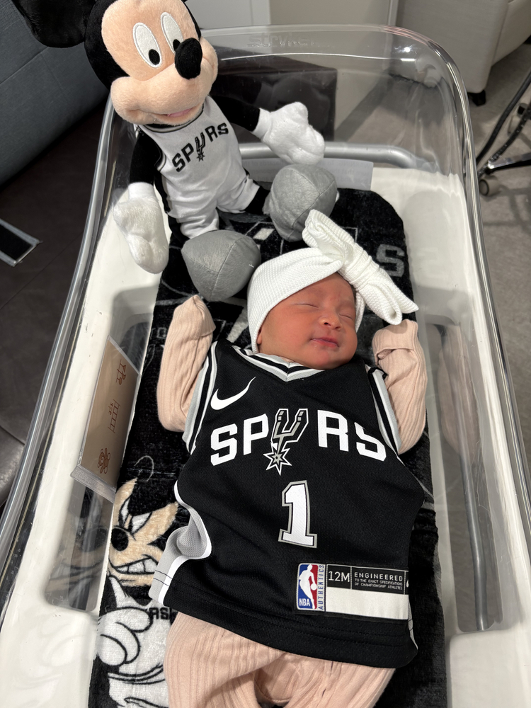 Newborn baby in hospital crib, wearing a spurs jersey and a spurs plush toy.