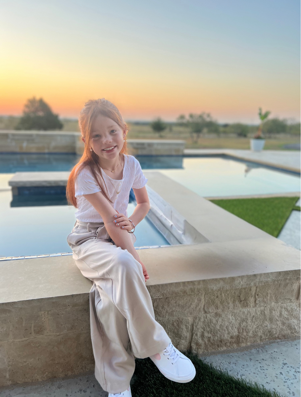 Young girl smiling and posing for a picture, while sitting next to a pool.