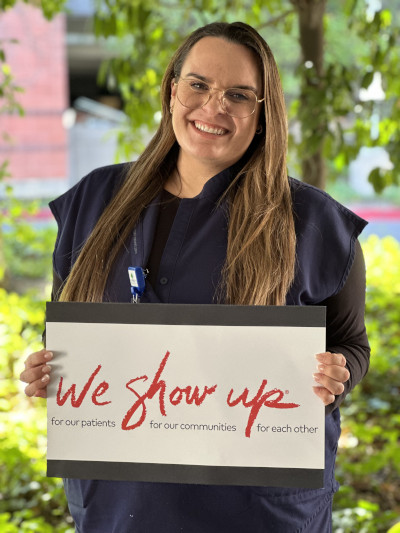 A nurse smiles while wearing navy blue scrubs and holding a sign that has the phrase, "We Show Up® for our patients. For our communities. For each other."