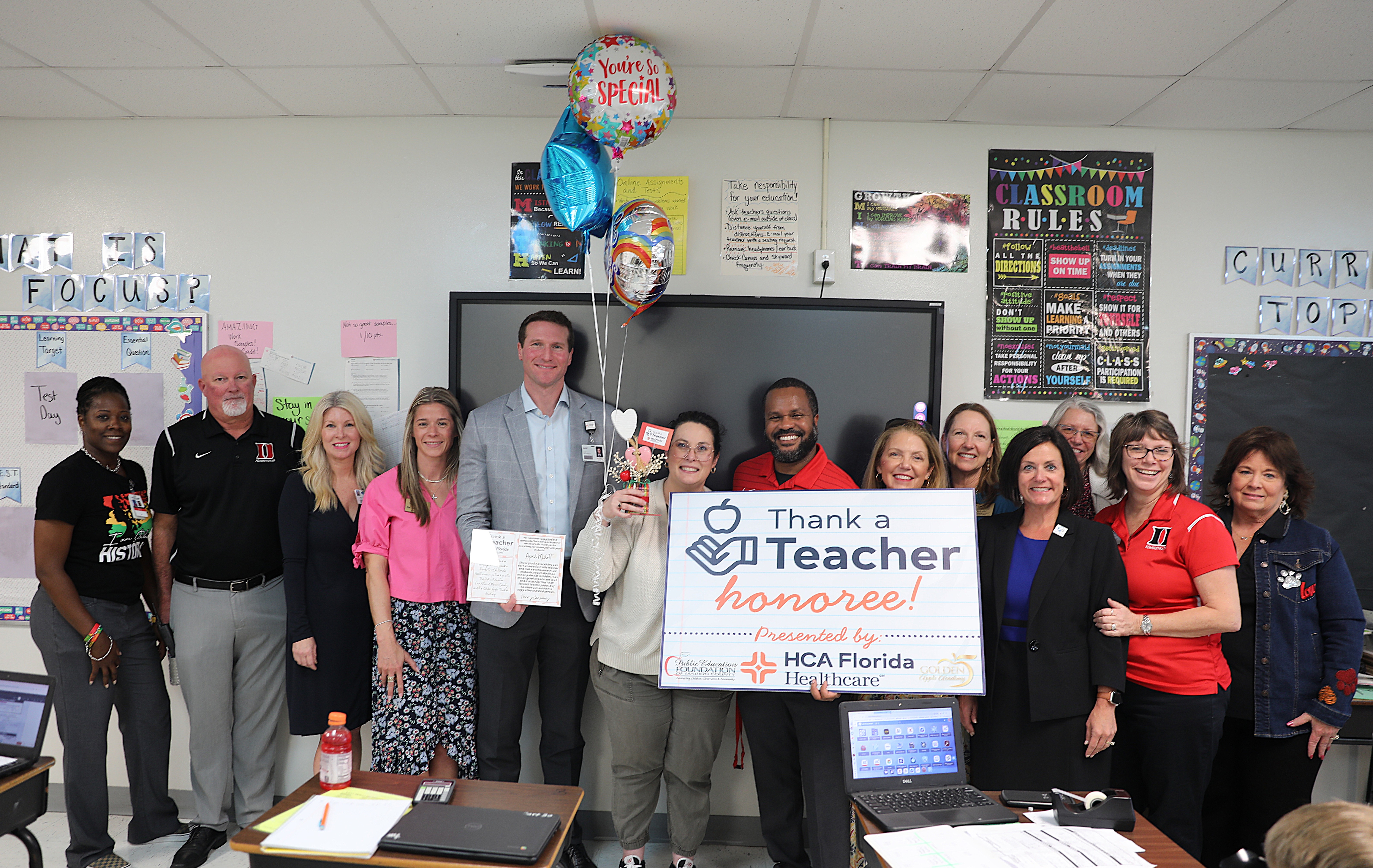 Group of school staff standing in a classroom celebrating a teacher who was recognized as a "Thank a Teacher Honoree" by HCA Florida Healthcare. The honoree stands in the center holding a certificate and small gift while colleagues surround her, and balloons float above them. A large sign reading "Thank a Teacher Honoree" is displayed in front of the group.