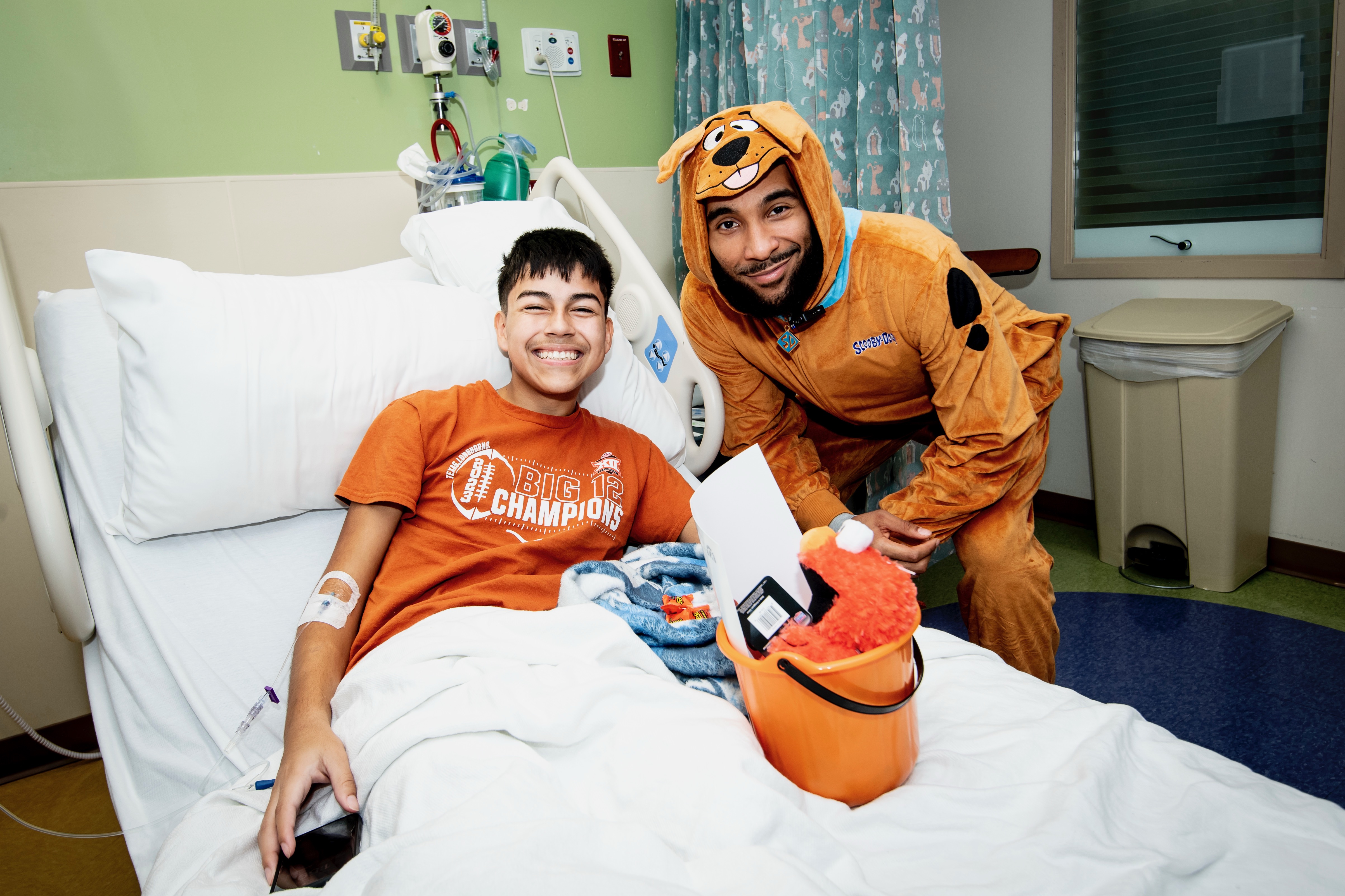 Julian Champagnie, San Antonio Spurs forward, hands out candy to a patient laying in a hospital bed while dressed as Scooby Doo.