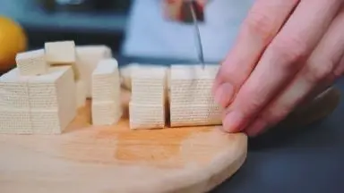 A person's hands holding and slicing chunks of tofu with a knife.