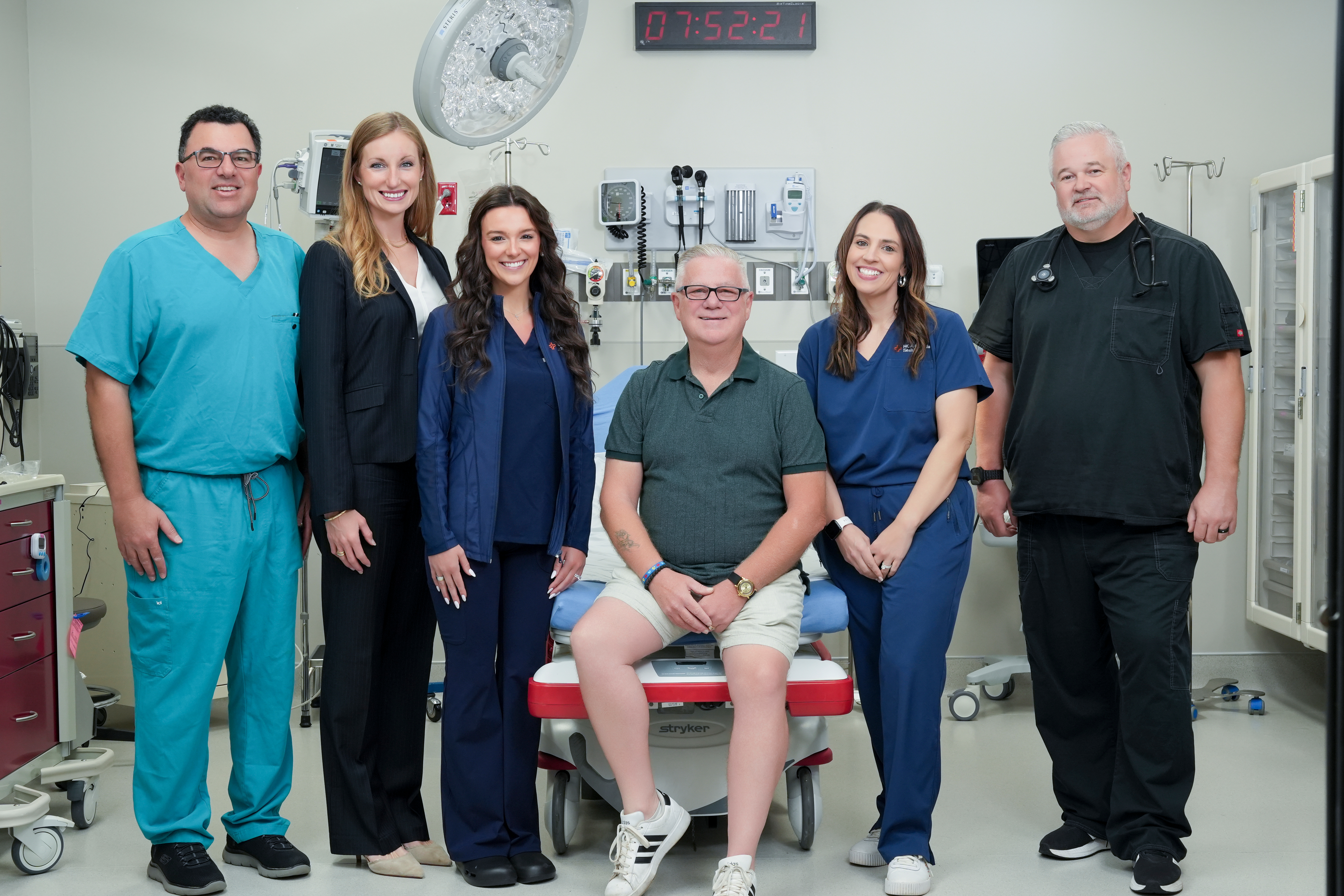 Darel Albin sits on an exam bed at HCA Florida Wellen Park Emergency, surrounded by the emergency care team, posing together in a hospital treatment room with medical equipment in the background.