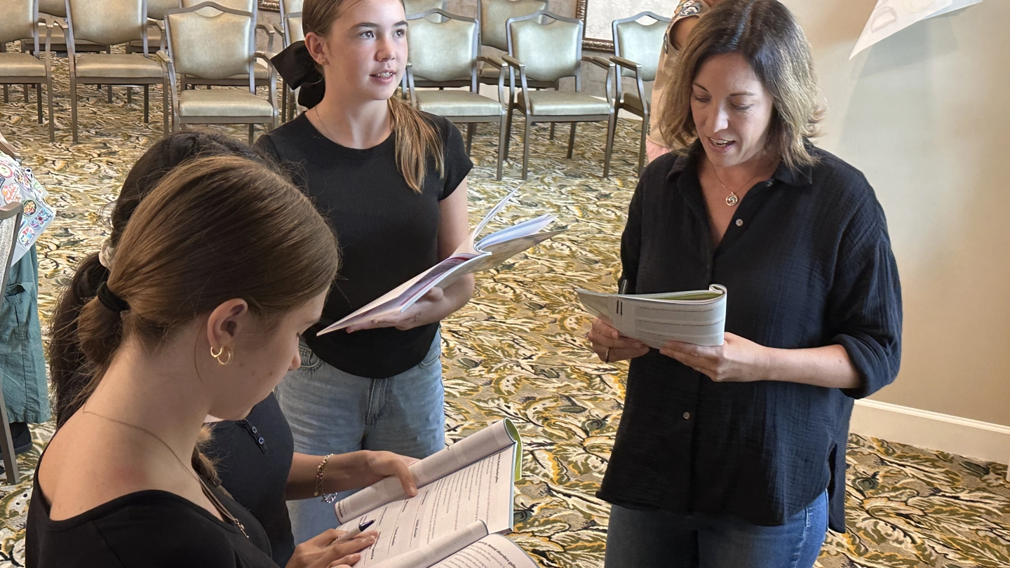 Two individuals stand side by side, writing in booklets during a Youth Mental Health First Aid training session. The indoor setting includes patterned carpet, chairs, tables and other participants engaged in activities.