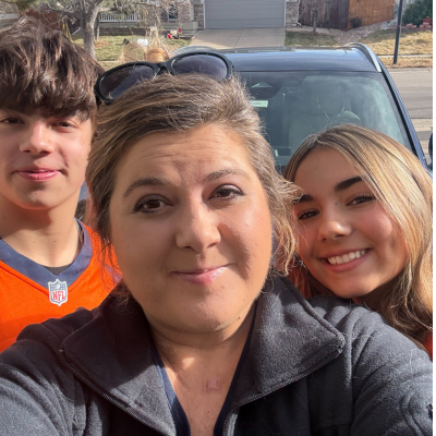 Three family members smiling for a selfie outdoors in front of a parked vehicle on a sunny day.