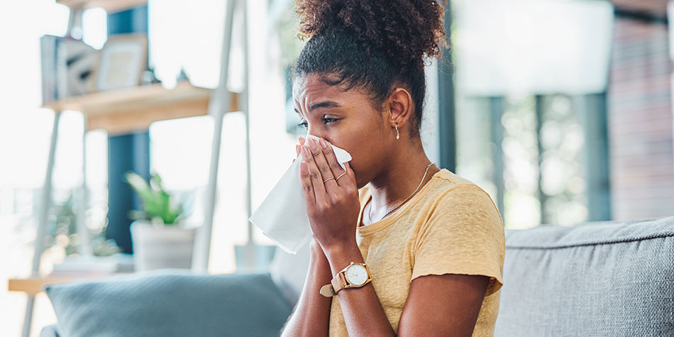 A woman blows her nose, while seated on a couch, with a set of shelves behind her in the background.