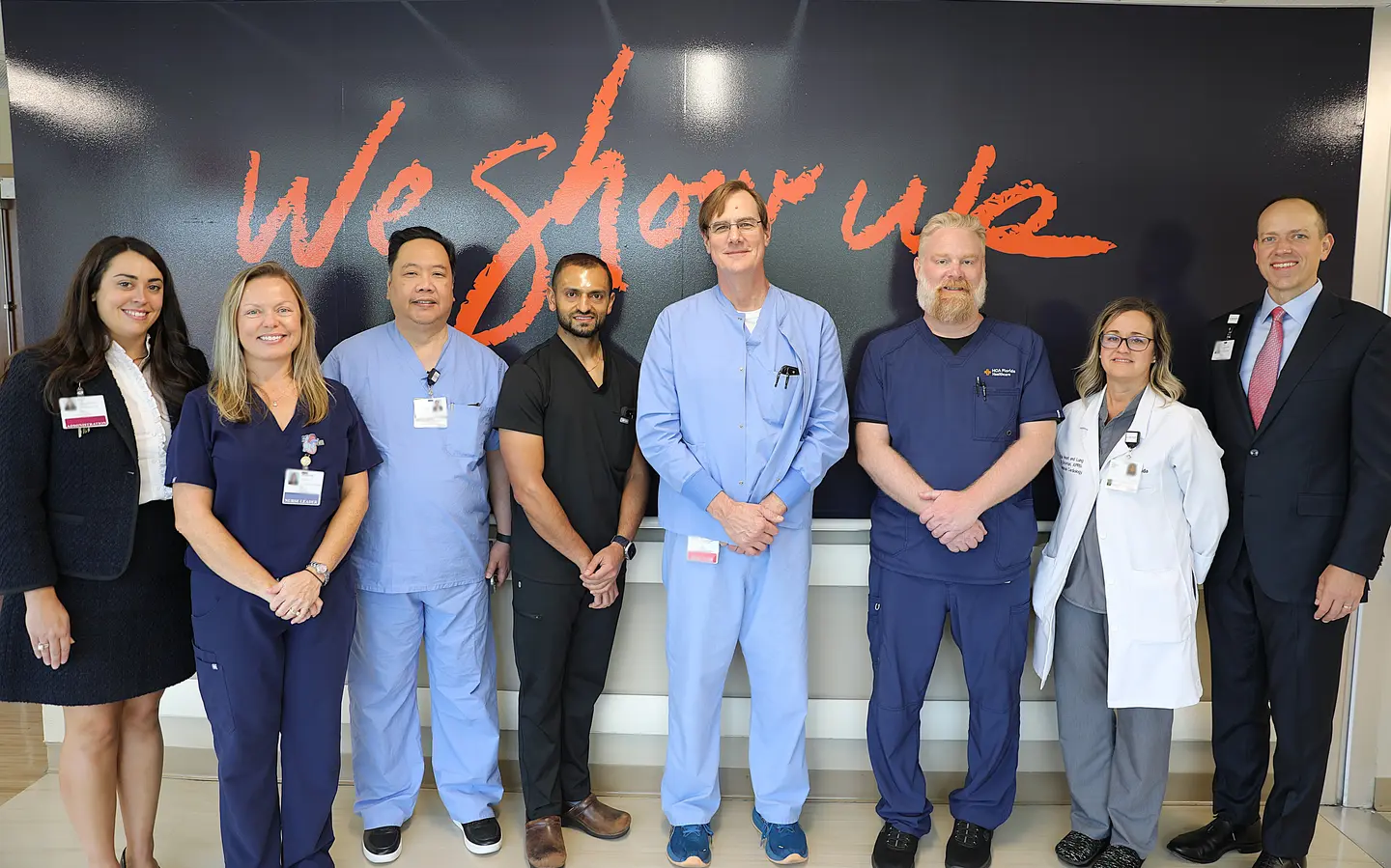 Five men and three women, some in suits and some in hospital scrubs standing in front of a large dark blue mural that says "We Show Up" in orange letters.