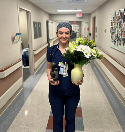 ICU RN Kelley Torrance holds award and bouquet of flowers in hospital hallway.