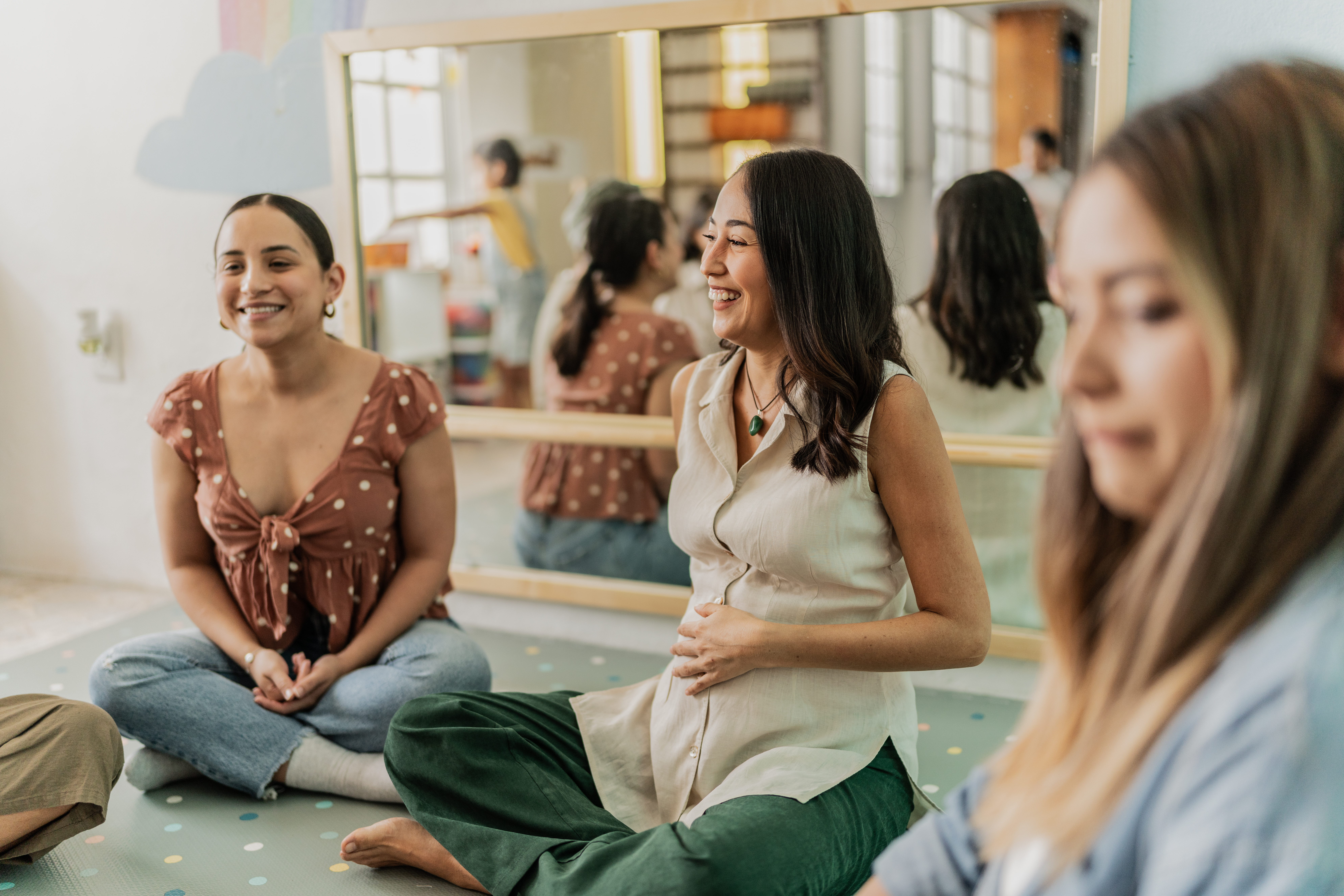 Three women sit on a mat for a prenatal class. One is out of focus in the foreground; the other two sit smiling at the center--one has her hand over her pregnant belly.