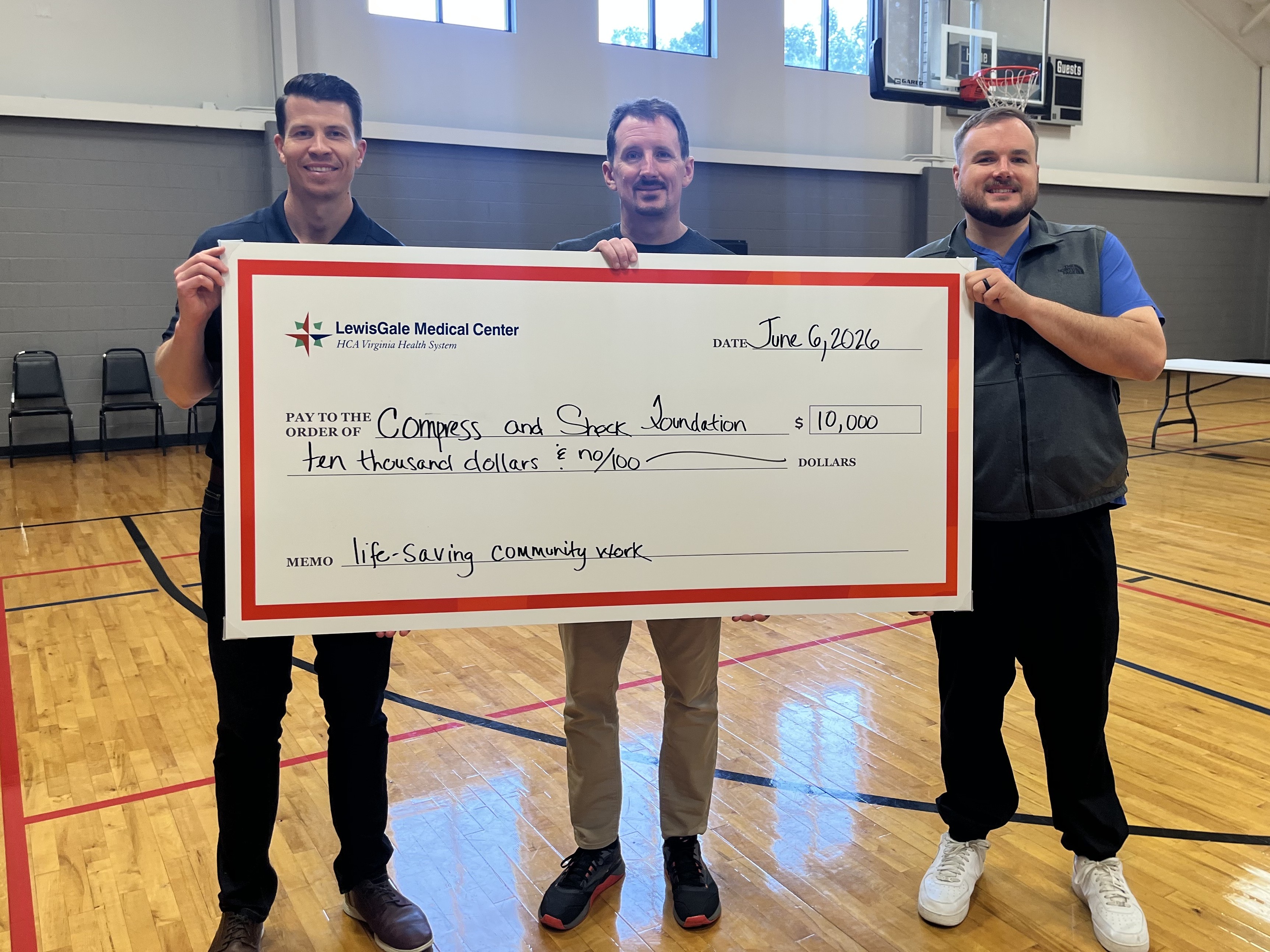 Three men stand in a gymnasium holding a large ceremonial check.