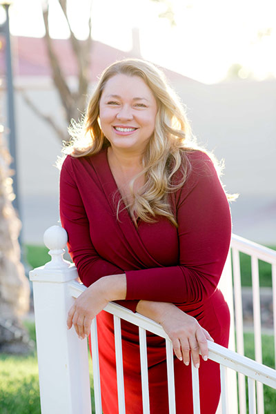 Alison Henriksen smiles while leaning against a railing with her arms crossed, wearing a red long-sleeved shirt and red skirt.