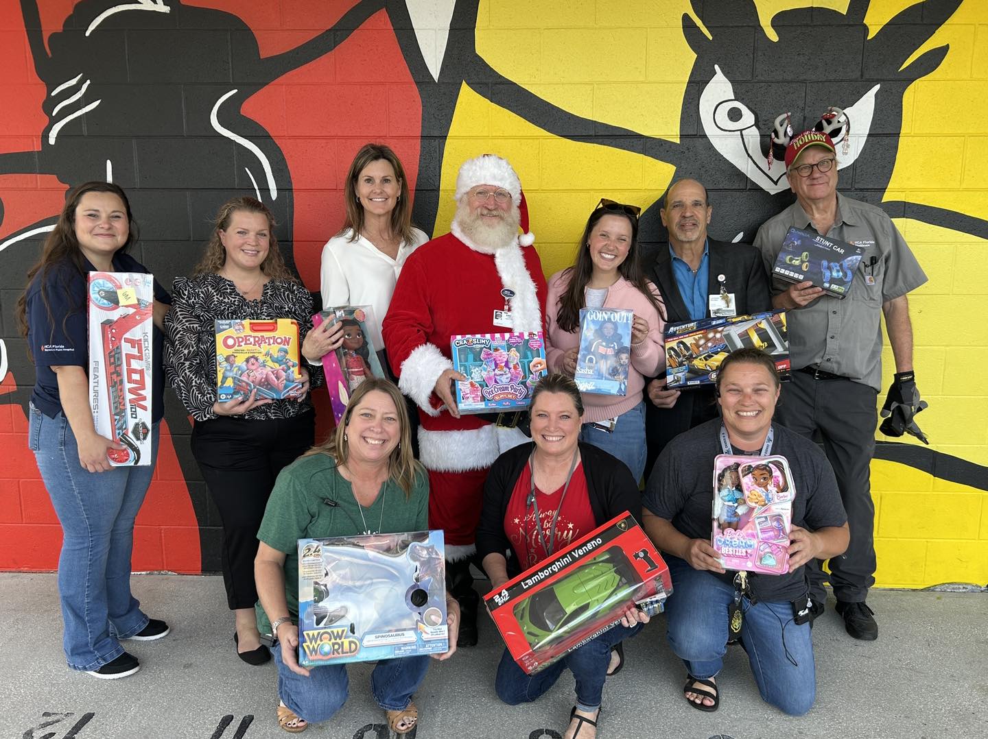 10 Hospital employees are standing in front of an art wall each holding a gift that will be donated to students at the school