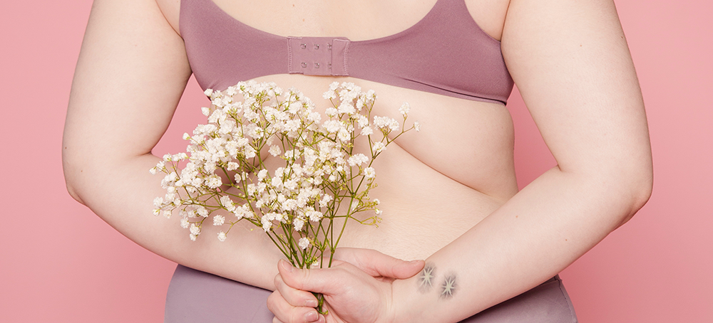 A woman, wearing a bra and stretch pants, holds a bouquet of Baby's Breath flowers behind her back.