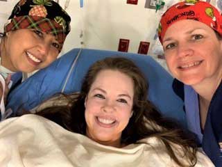 Nurse Stacy Ciaschini in her hospital bed smiling, with two of her nurse colleagues at her bedside.