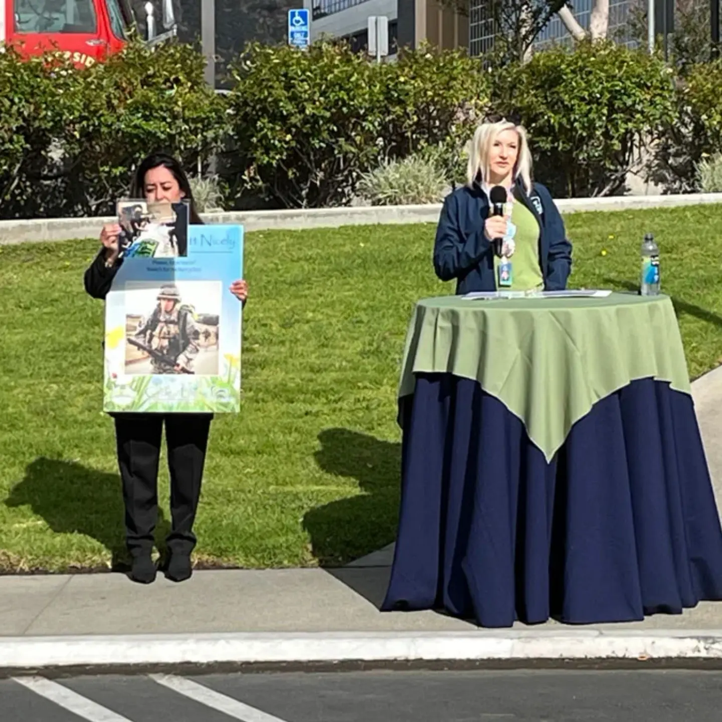 Nicole Heimerl speaks at a standing table about her son who lost his life in a motorcycle accident. An assistant holds up a poster and pictures of Austin.