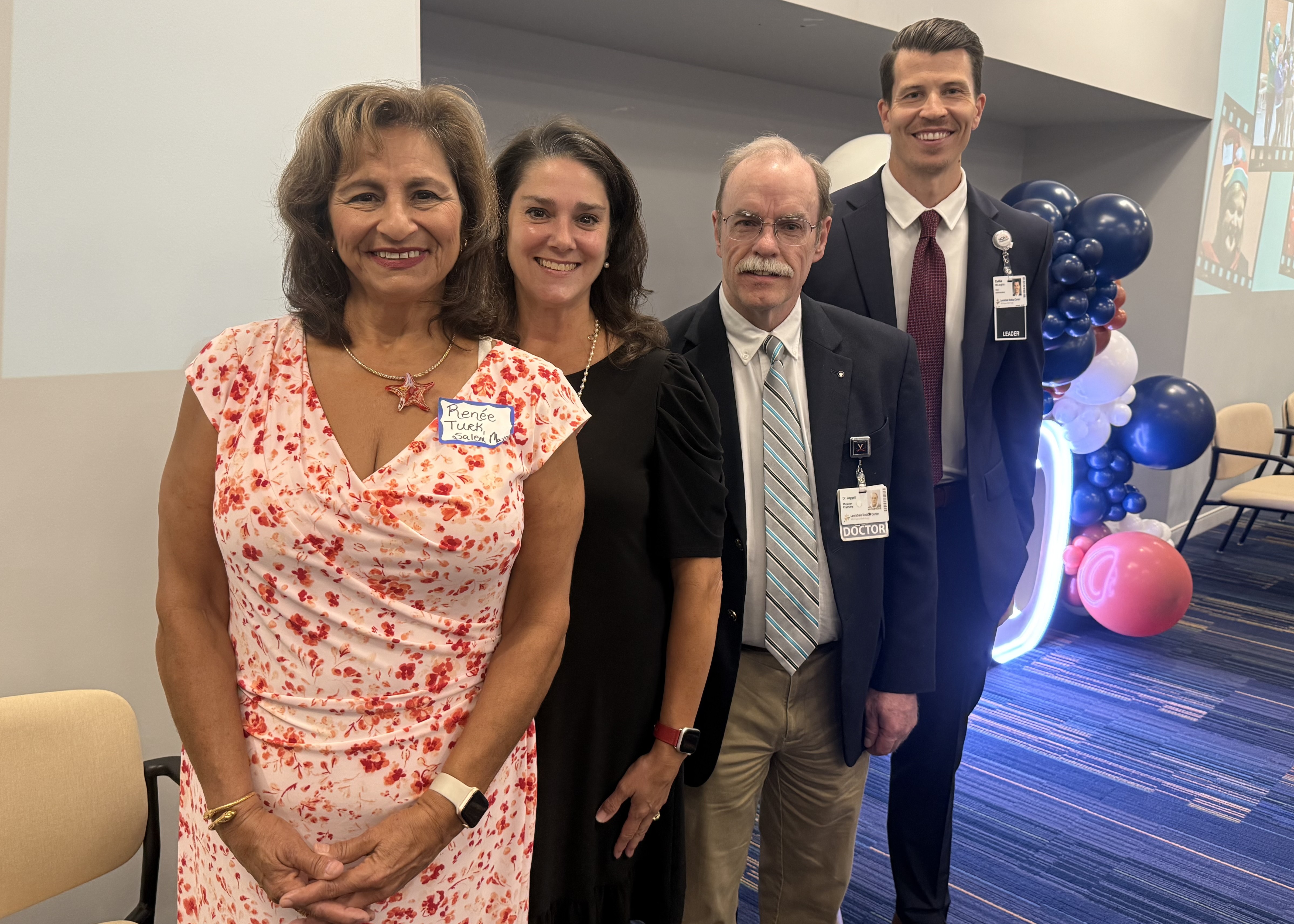 Four people stand in a row. Two women wearing dresses and two men wearing suits. There are balloons behind them. They are each smiling.
