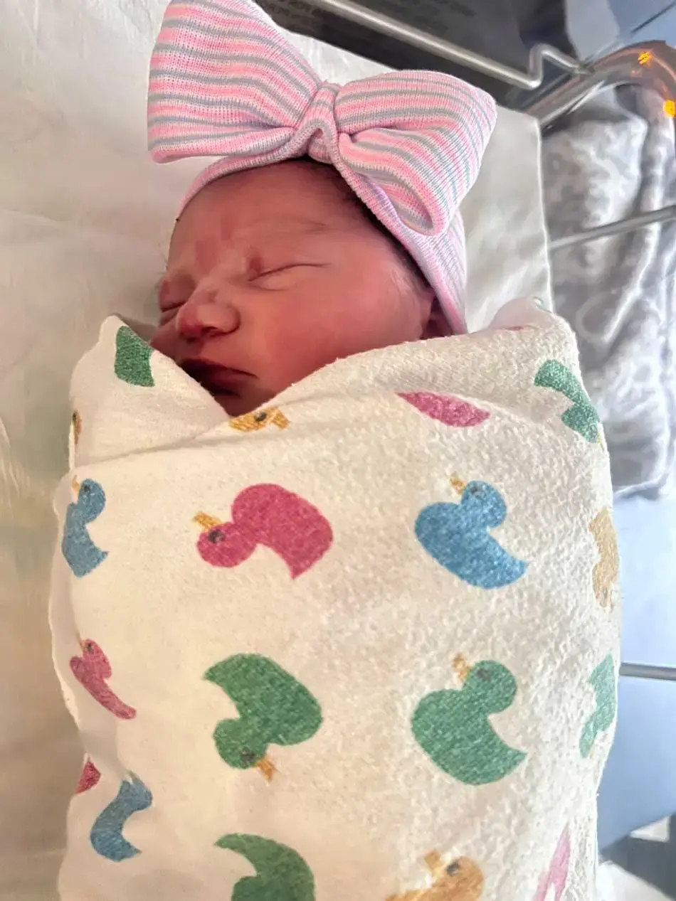 Baby Lozano sleeps in a crib, wrapped in a white blanket with multicolored ducks on it and wearing a pink and gray hat with a bow.
