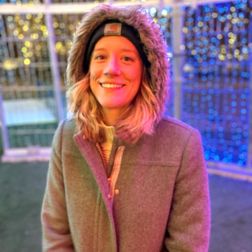 Megan Titcomb smiles while standing under a winter light display, wearing a hooded jacket and a beanie hat.