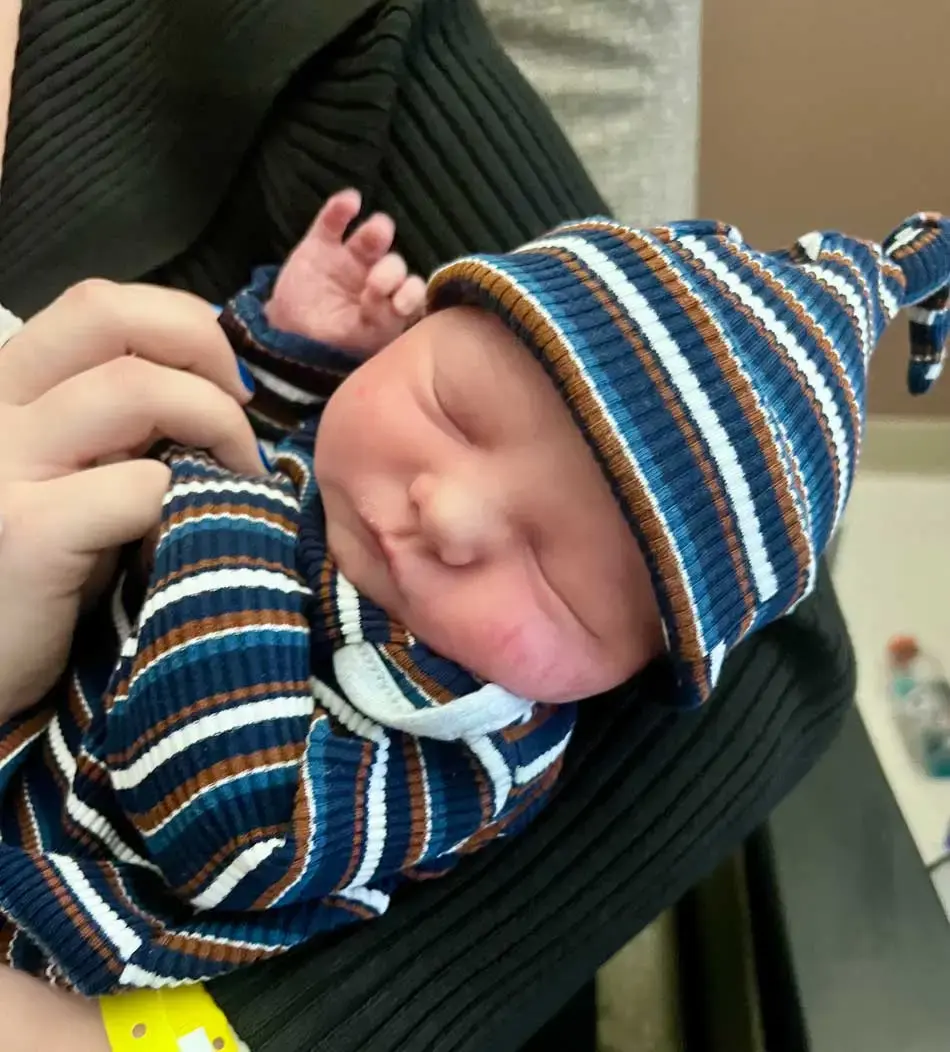Baby William sleeps while being held by his mother Taylor, wearing a blue, brown and white striped outfit.