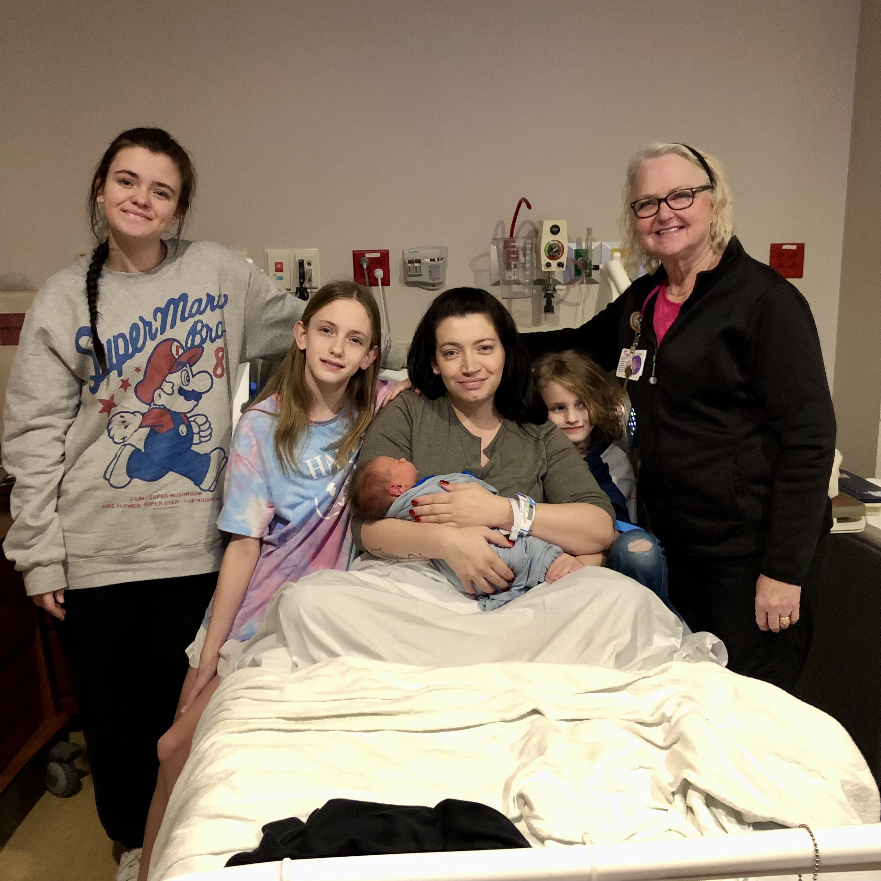 Three children and a female lactation consultant stand beside the hospital bed with a mother holding a new baby brother. -Elizabeth (15), Madelynn (11), Sarah (mother) holding Barrett, Rowan (6), and Joy (lactation consultant)