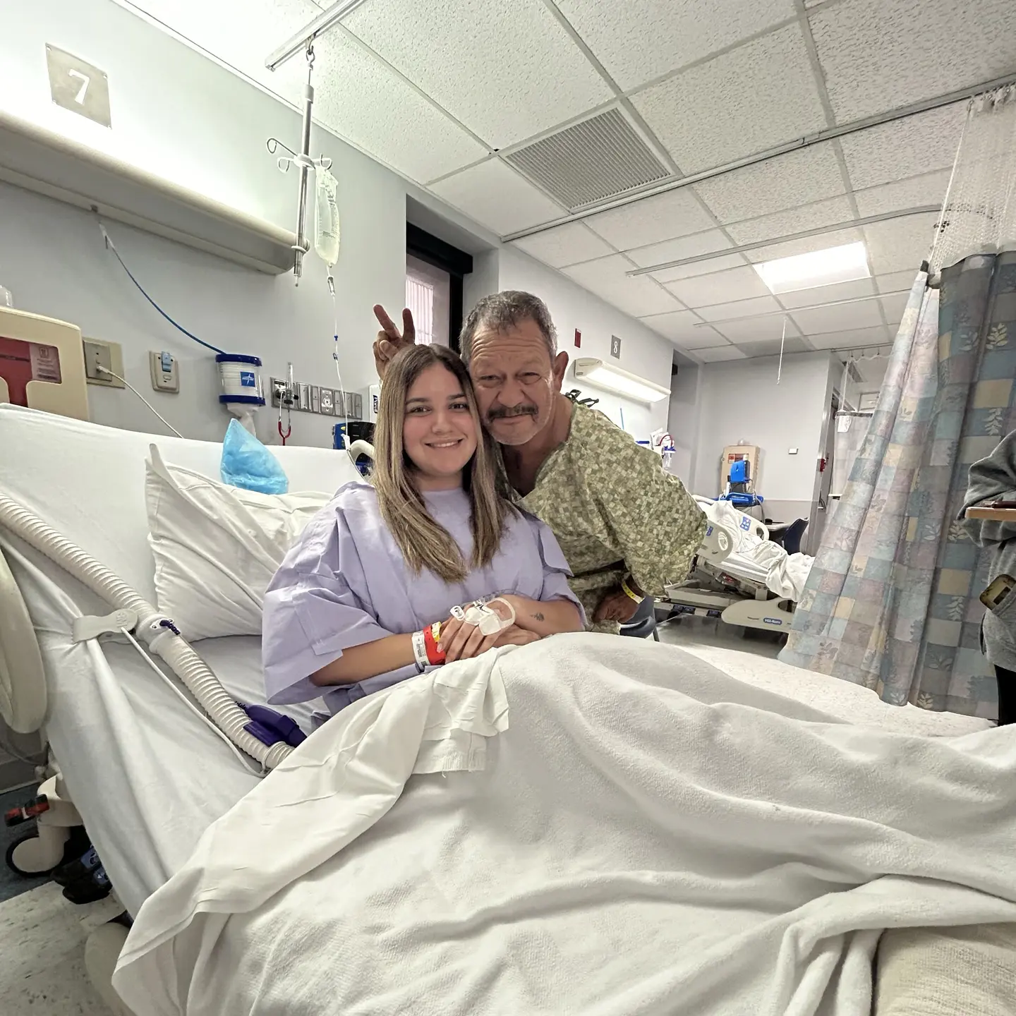 Itzel Guzman posing with her father (giving bunny ears/peace sign above her head) in hospital surgery bed