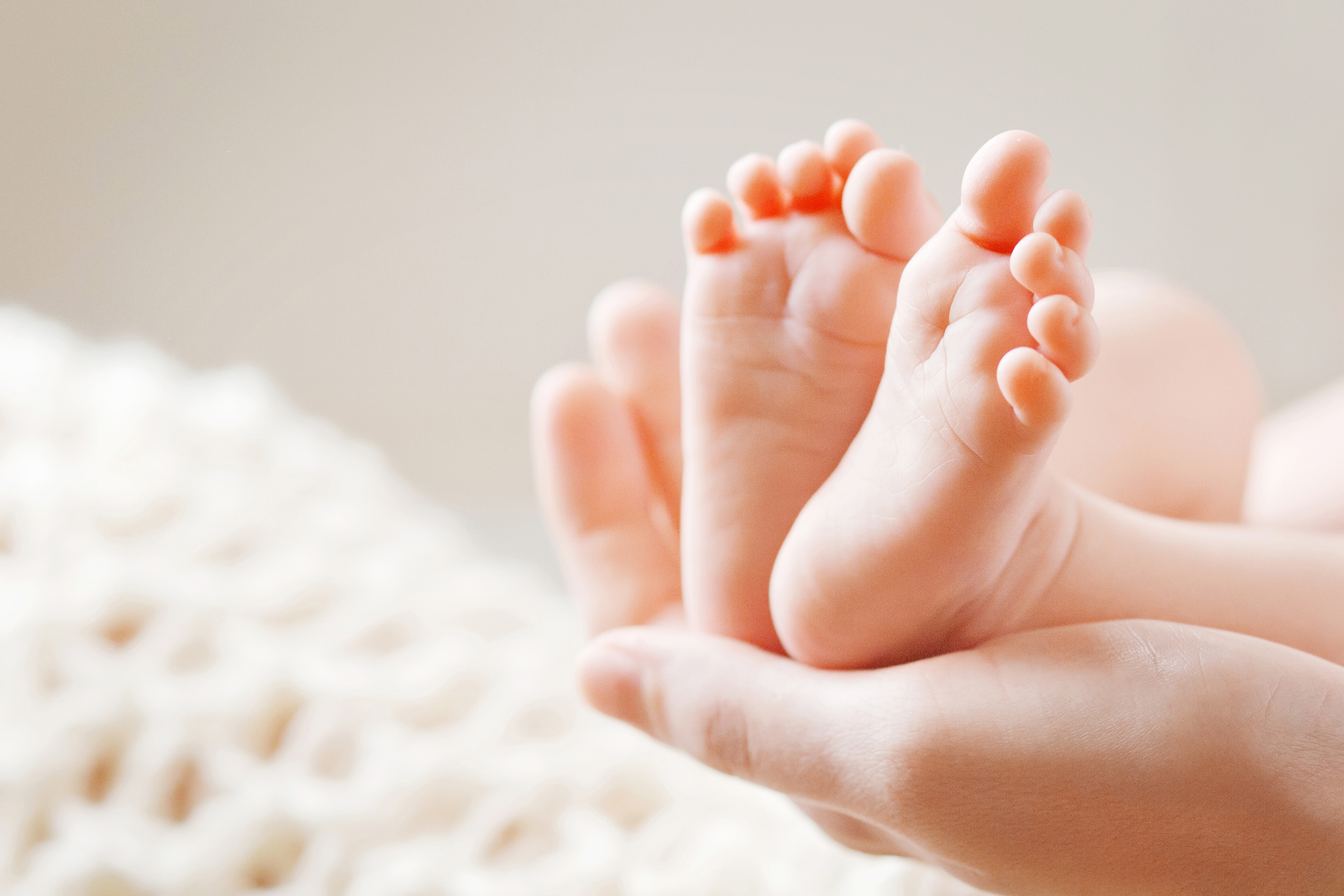 Adult hands holding the tiny feet of a newborn baby, with a soft blanket in the background.